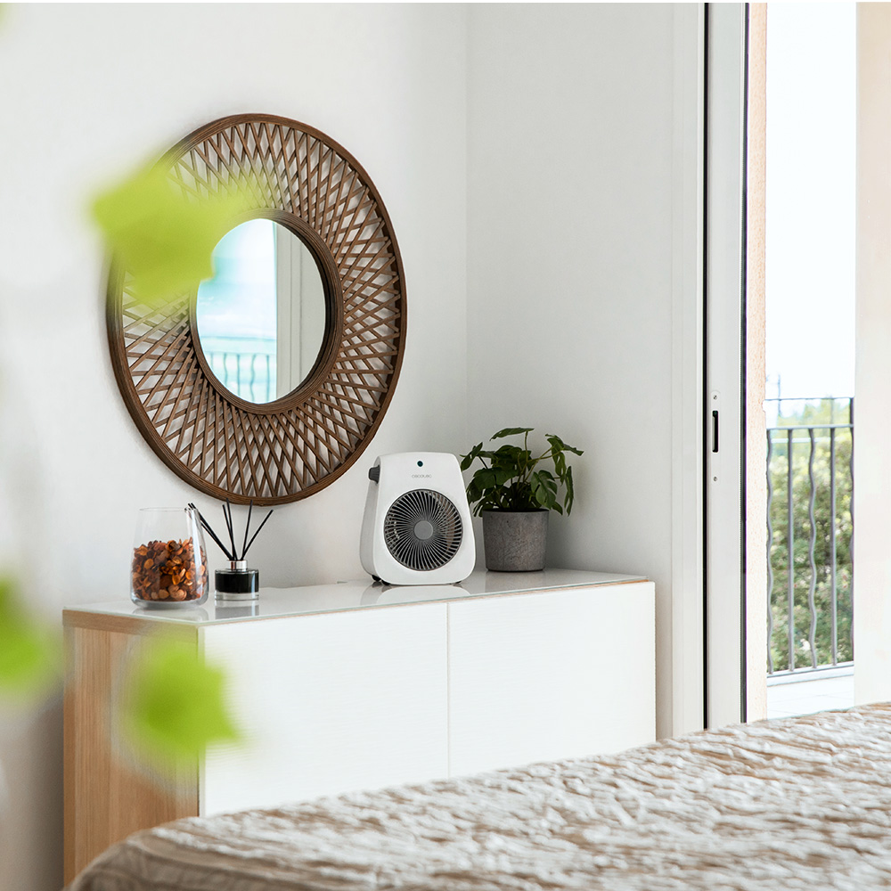 Un ventilador blanco, un espejo y una planta en maceta están sobre un mueble blanco. Por la puerta abierta, un balcón es visible.