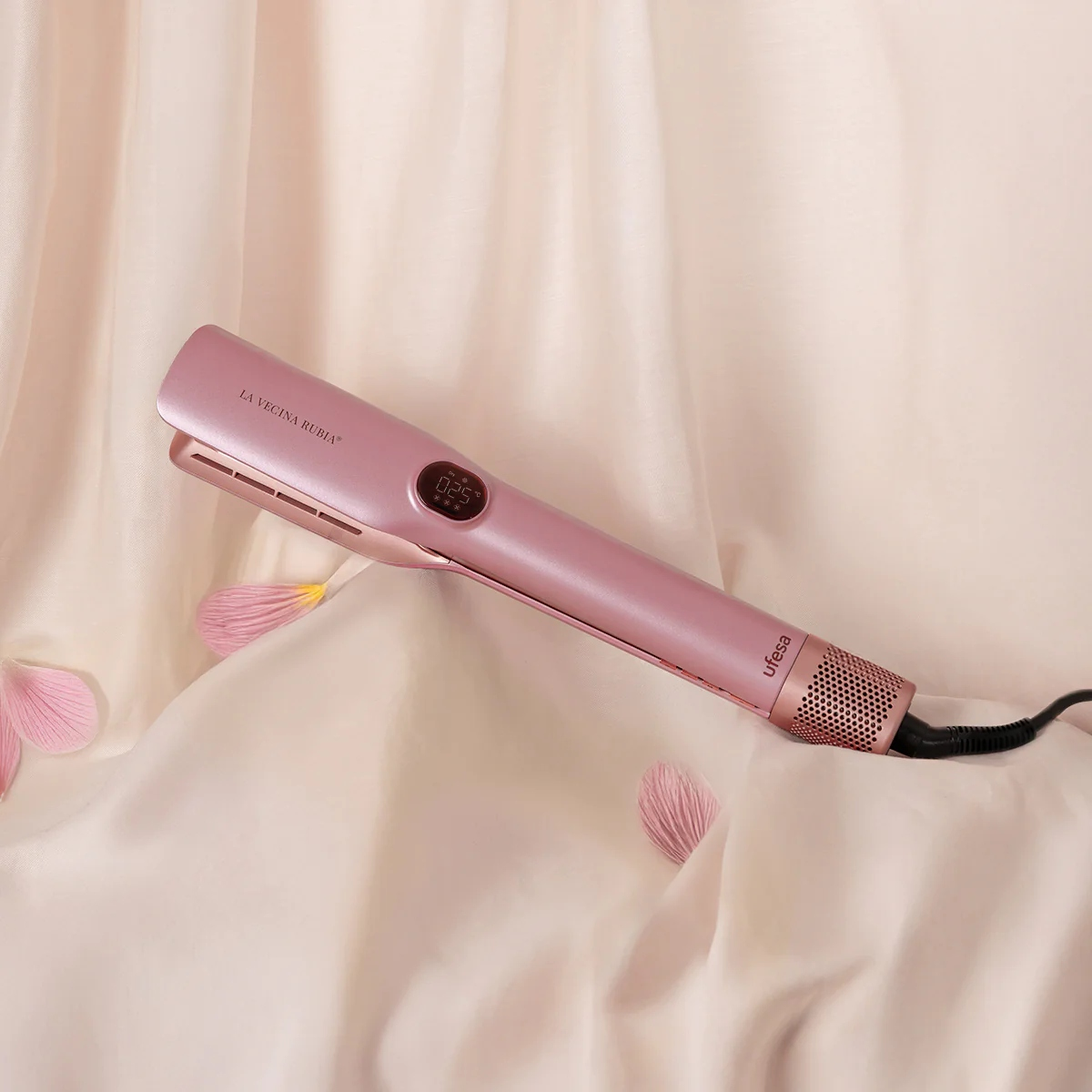 Una plancha de pelo rosa sobre un paño crema con pétalos rosas. La plancha tiene una pantalla digital y el nombre de la marca.