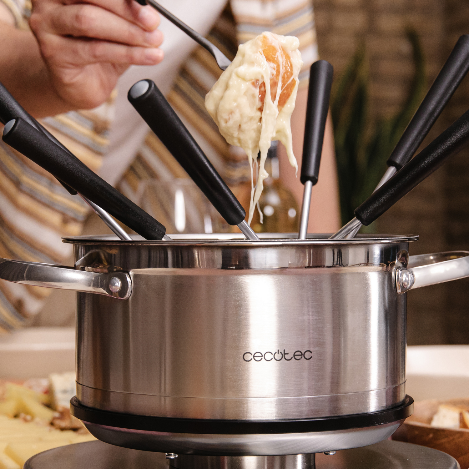 Una olla de fondue con tenedores, queso y una persona sumergiendo comida. La olla es de acero inoxidable. Se está sumergiendo queso.