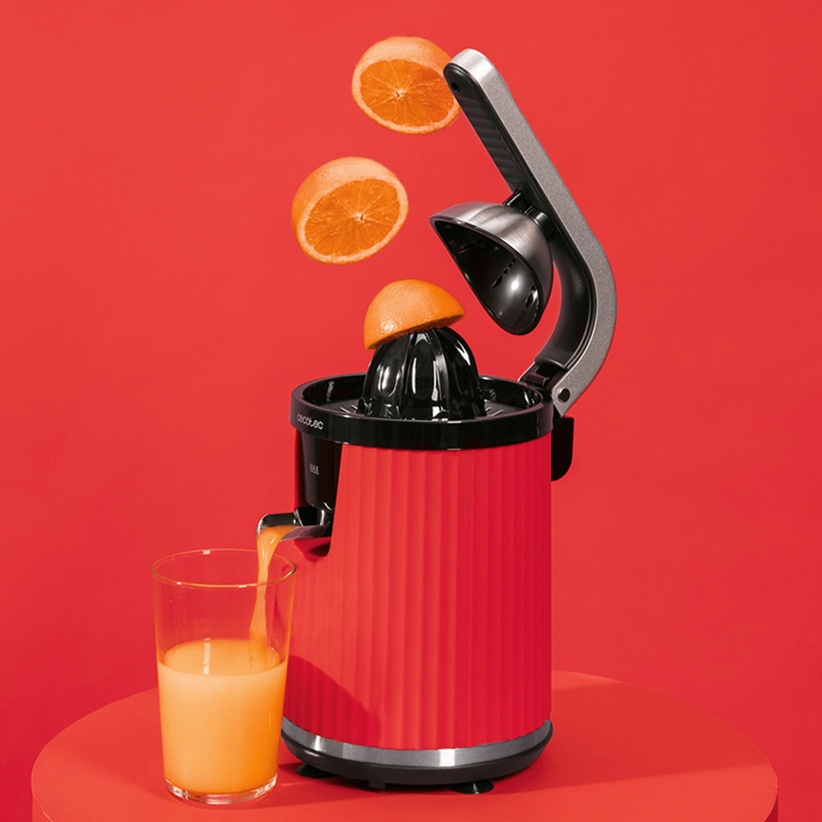 Una exprimidora roja con media naranja encima, naranjas flotando arriba y jugo vertiéndose en un vaso sobre un fondo rojo.