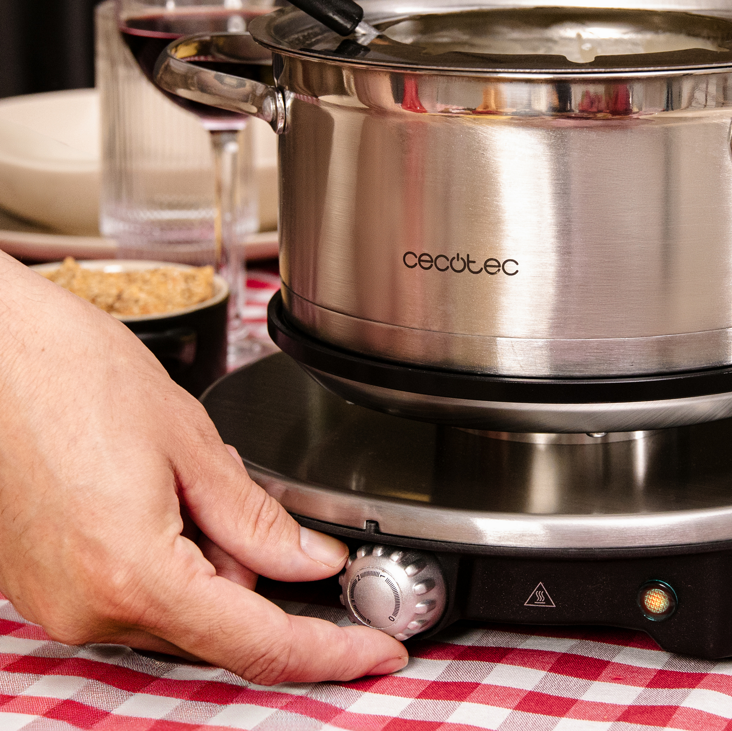Una mano gira un dial en una olla de fondue. Una olla de acero inoxidable encima. Mantel a cuadros rojos en primer plano.