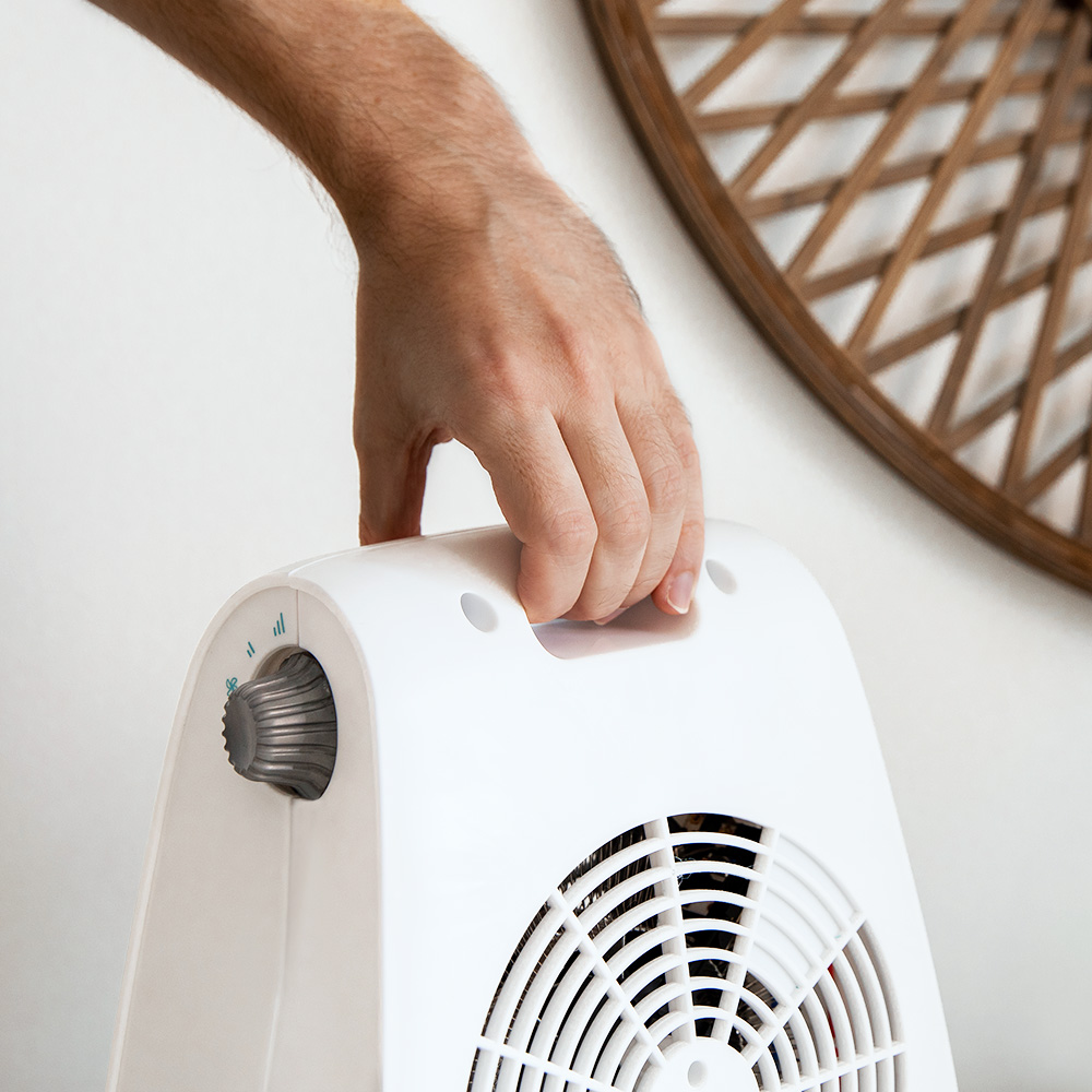 Una mano sostiene un ventilador blanco. En el fondo, un elemento decorativo de pared de madera. El ventilador tiene una perilla y una rejilla negra.