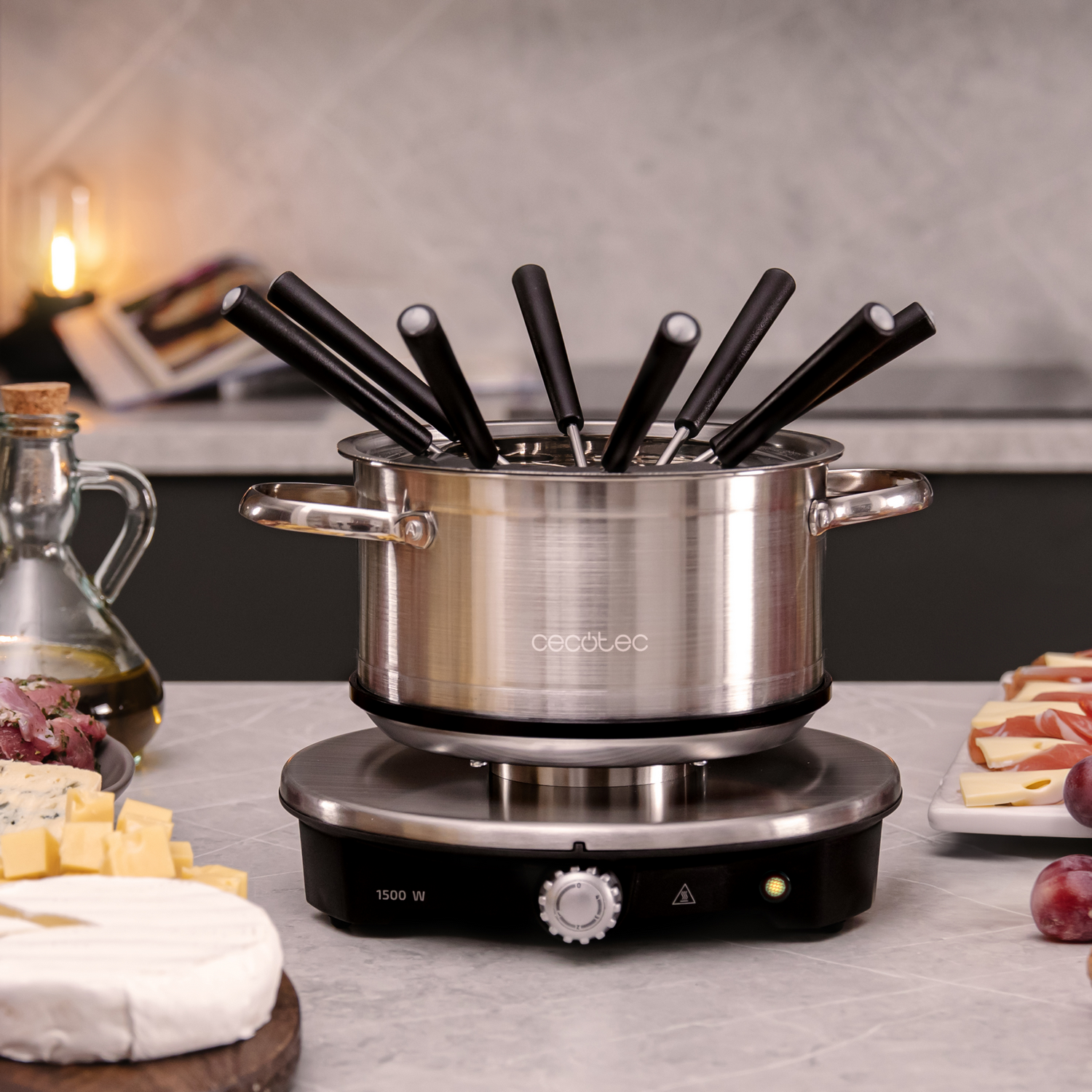Una olla de fondue de acero inoxidable con tenedores con mango negro. Queso, carne y uvas la rodean sobre mármol.