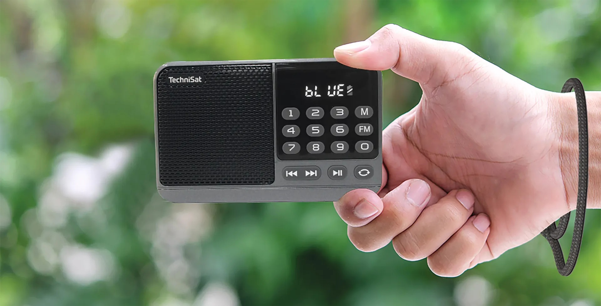 Dłoń trzyma szare radio TechniSat, wyświetlające na ekranie 'BLUE'. Czarna bransoletka na nadgarstku. Rozmyte zielone tło.