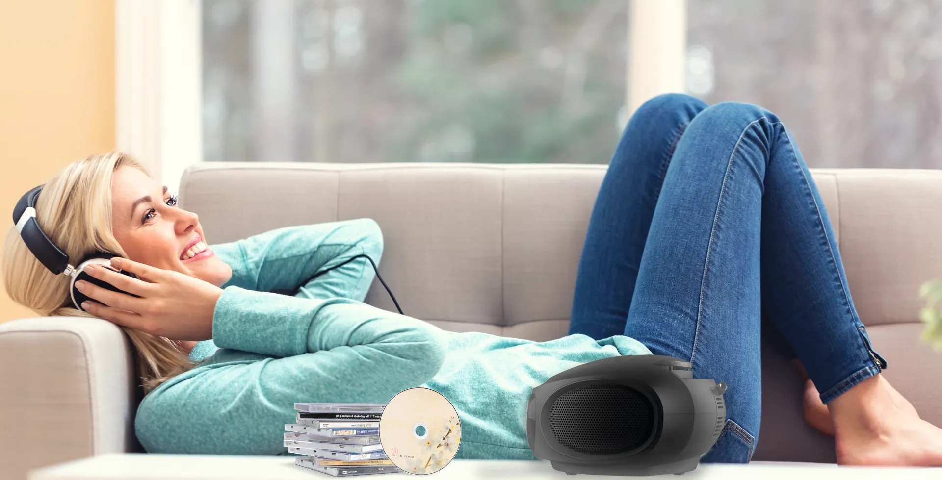 Kobieta na kanapie w słuchawkach. Płyty CD i głośnik przed nią. Uśmiecha się i słucha muzyki.
