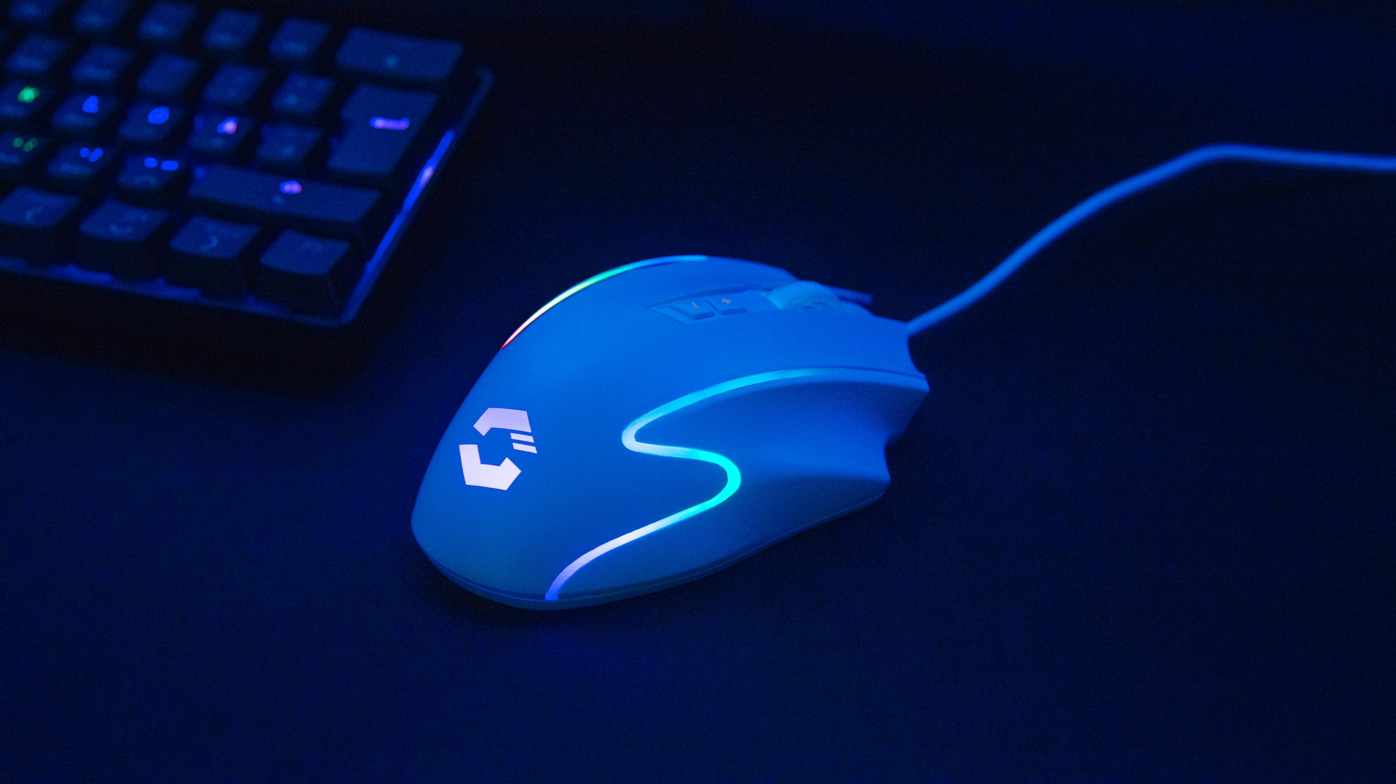 Eine weiße Gaming-Maus mit leuchtendem Logo und Lichtern liegt auf einer dunklen Oberfläche, im Hintergrund eine Tastatur.