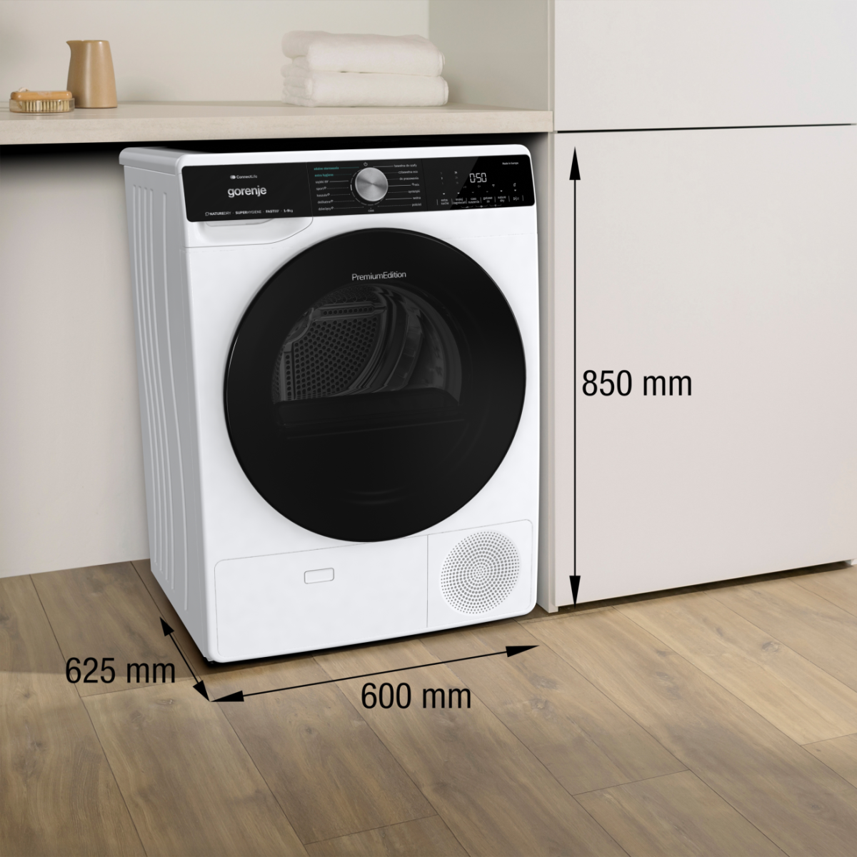 Biała suszarka Gorenje z czarnymi drzwiami. Wymiary: 600mm x 625mm, wysokość 850mm. Ręczniki i dzbanek w tle.