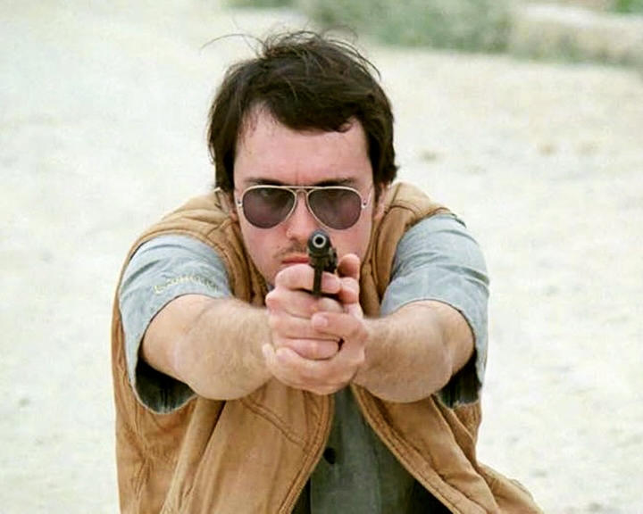 Ein Mann mit Sonnenbrille und brauner Weste zielt mit einer Pistole direkt auf die Kamera. Der Hintergrund ist eine Wüstenlandschaft.