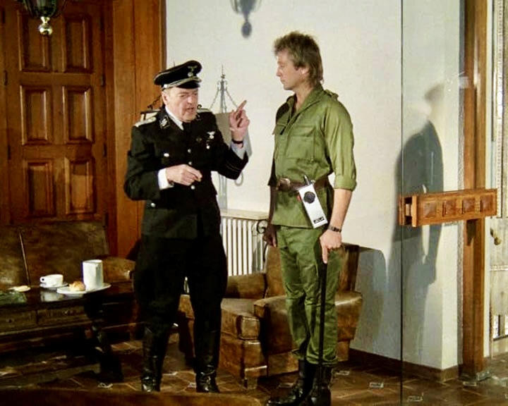Zwei Männer in einem Raum. Einer in Uniform zeigt. Der andere in Grün schaut zu. Holztür und Möbel im Hintergrund.