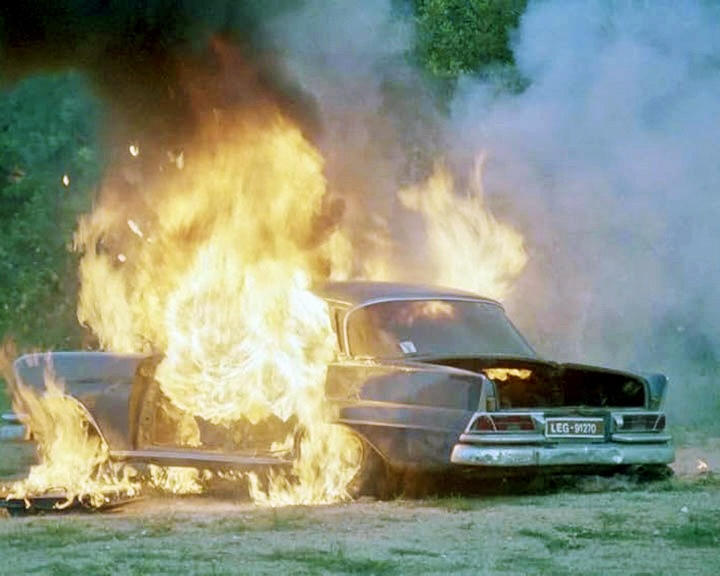 Ein Oldtimer steht in Flammen. Intensives orangefarbenes Feuer umgibt das dunkelfarbene Fahrzeug, Rauch steigt vor grünem Hintergrund auf.