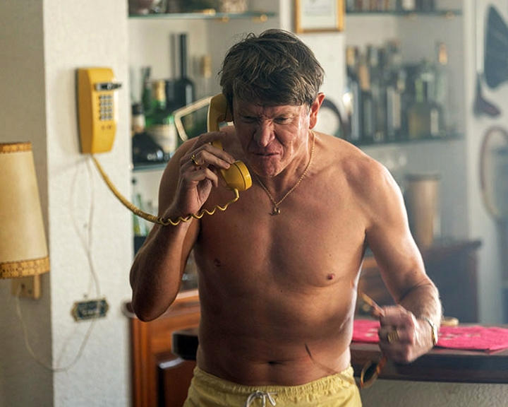 Oberkörperfreier Mann telefoniert, gelbe Shorts. Gelbes Wandtelefon, Lampe, Bar im Hintergrund.