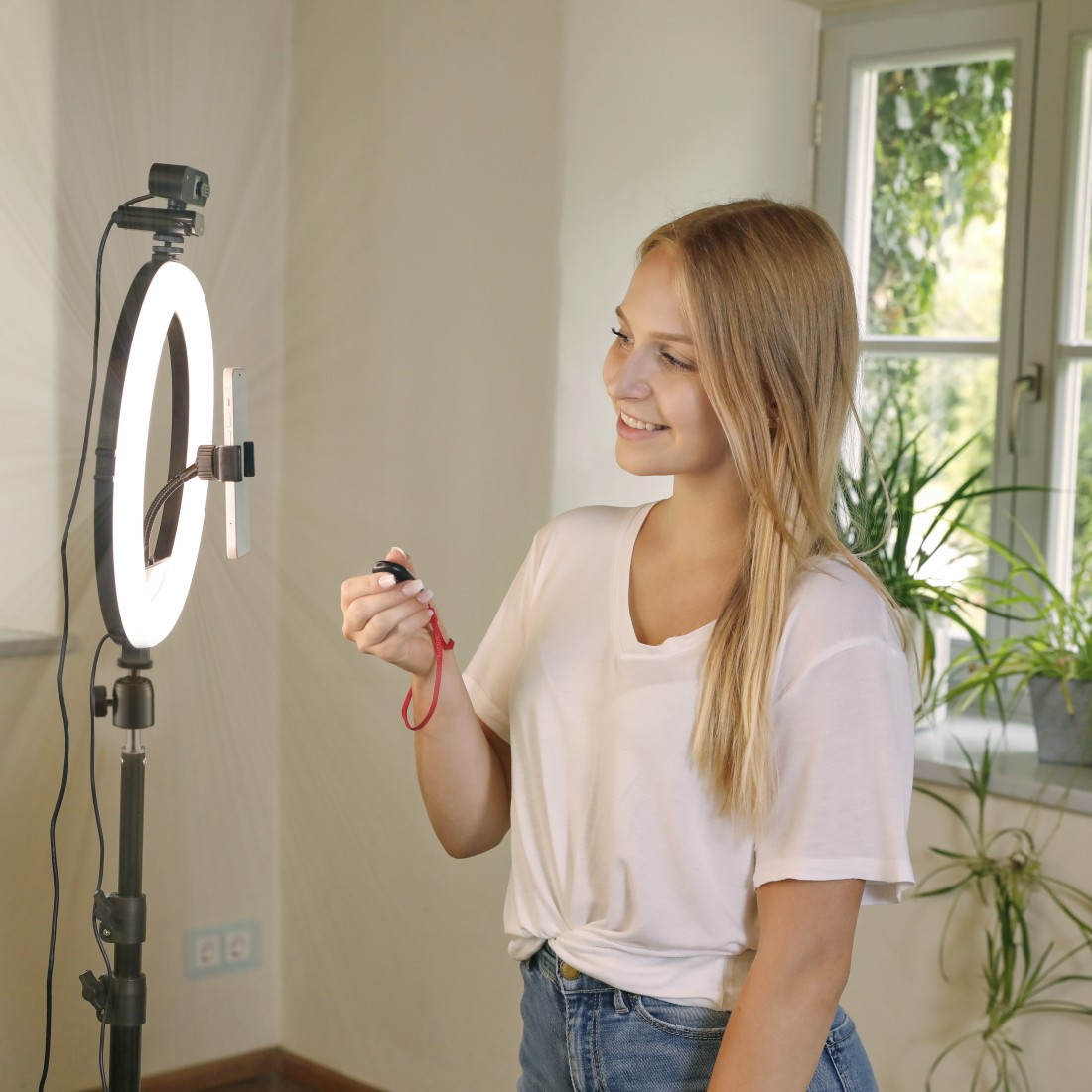 Eine lächelnde Frau hält eine Fernbedienung. Ein Ringlicht und ein Telefonständer sind auf sie gerichtet. Im Hintergrund ein Fenster mit Pflanzen.
