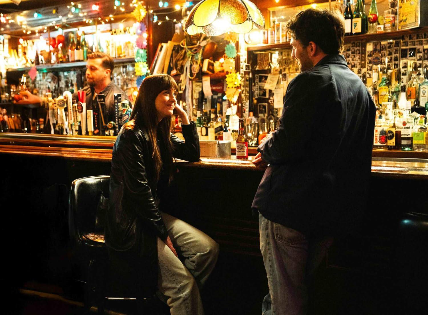 Eine Frau in einer Lederjacke sitzt an einer Bar und unterhält sich mit einem Mann. Ein Barkeeper steht hinter der Bar. Flaschen säumen Regale.