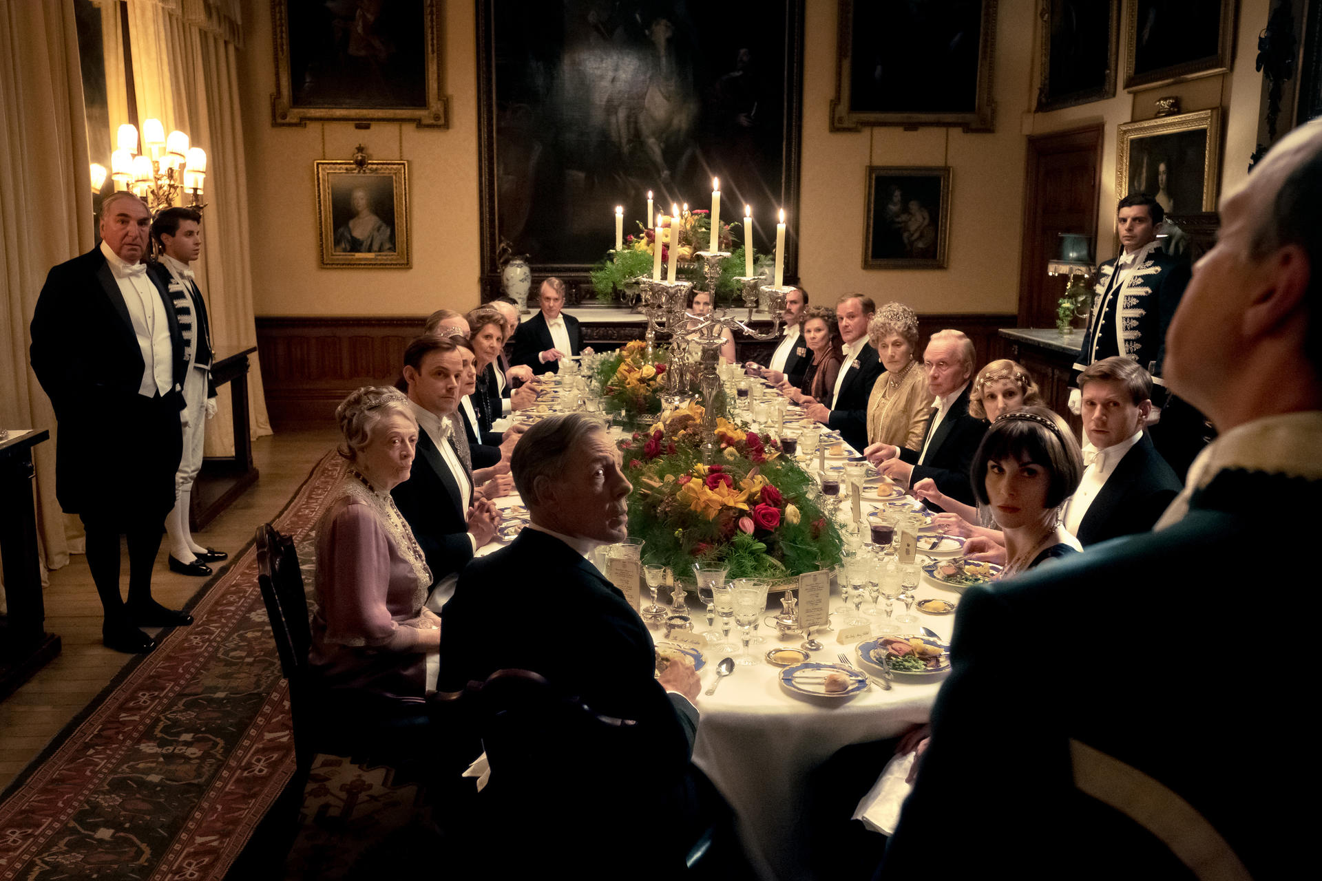 Formelle Dinnerszene. Personen sitzen an einer langen Tafel. Kerzen und Blumenschmuck zieren den Tisch. Kellner in Uniform stehen daneben.