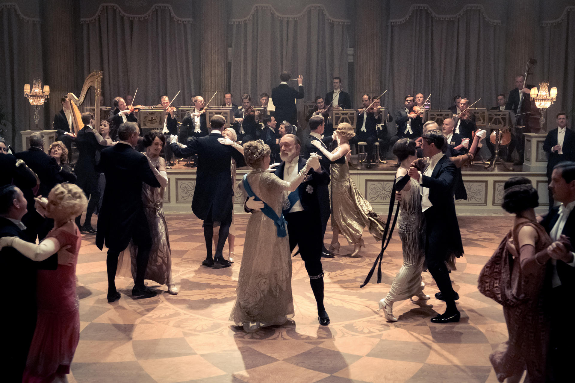 Eine Ballszene mit tanzenden Paaren. Im Hintergrund spielt ein Orchester. Frauen tragen Kleider, Männer Anzüge. Der Boden ist gemustert.