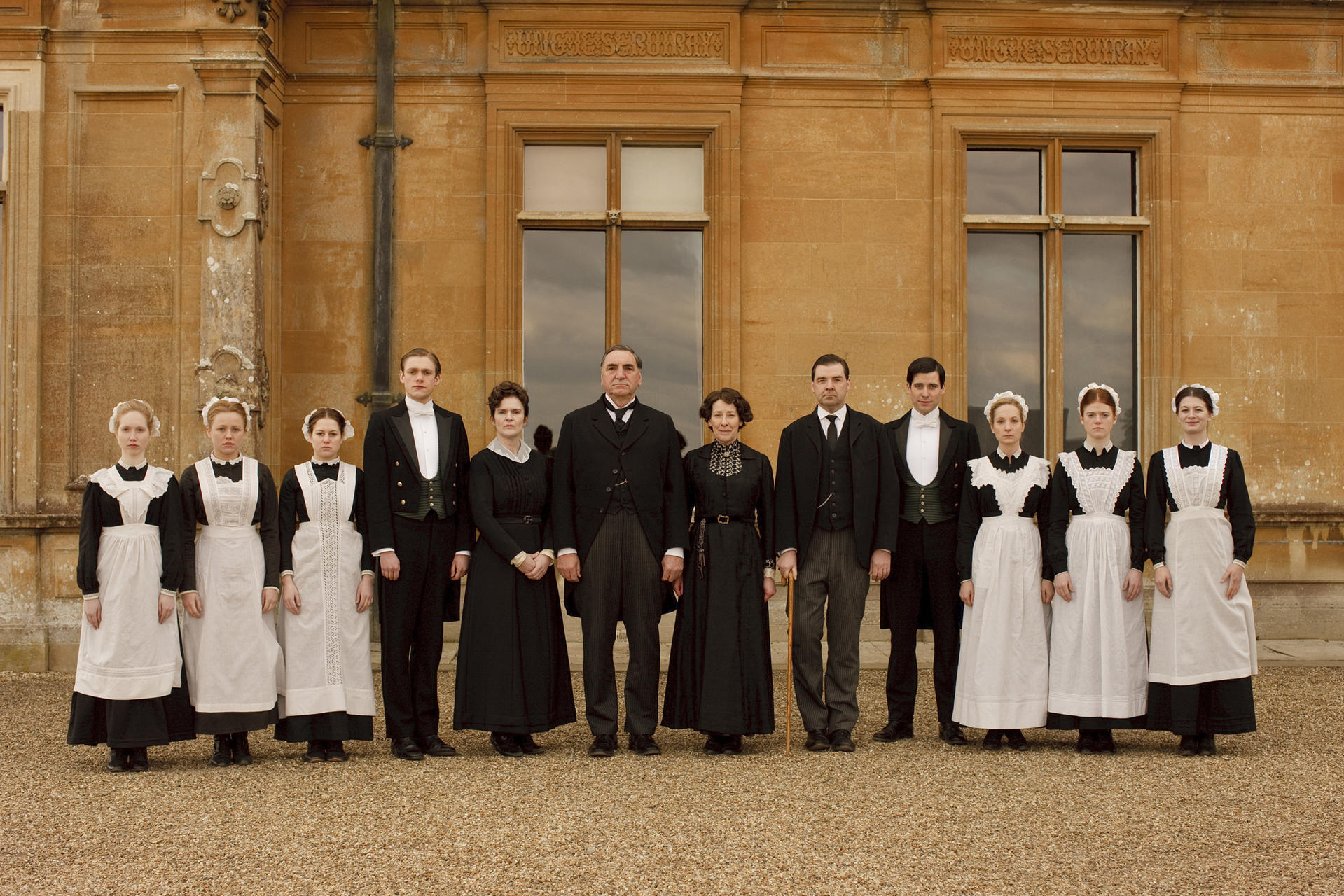 Eine Gruppe von Menschen in formeller Kleidung steht vor einem großen Steingebäude. Einige tragen Dienstmädchen- und Butleruniformen.