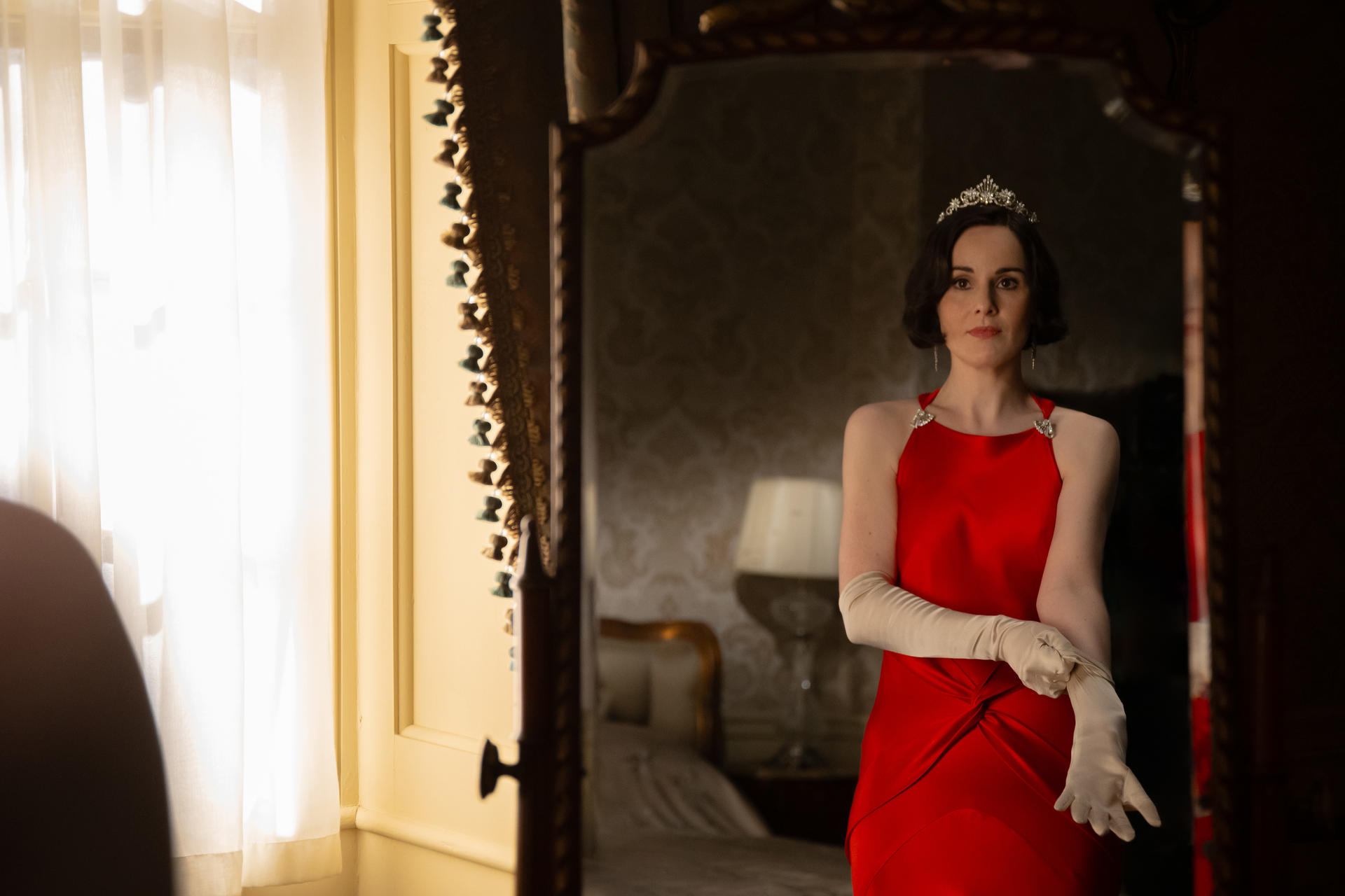 Frau in rotem Kleid, Diadem und Handschuhen, blickt in den Spiegel. Raum mit Fenster und Lampe im Hintergrund.
