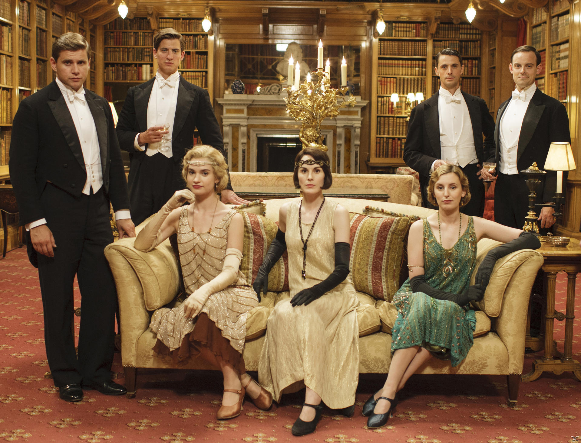 Formelles Gruppenporträt: Männer in Smokings, Frauen in Flapper-Kleidern, sitzen auf einem Sofa. Hintergrund: Bücherregale und Kamin.