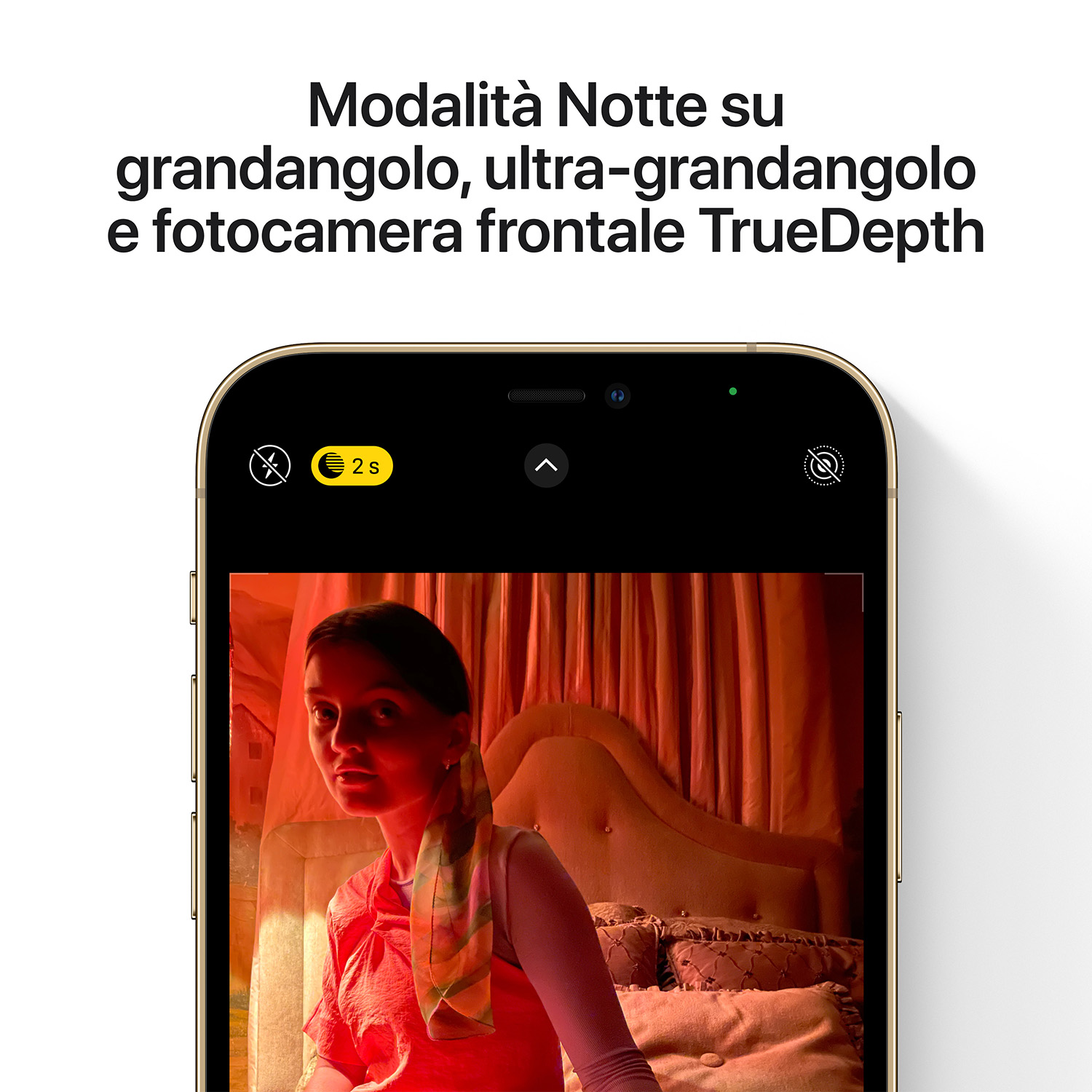 Una donna guarda la fotocamera. Il telefono mostra un'immagine in modalità notturna con un letto sullo sfondo.