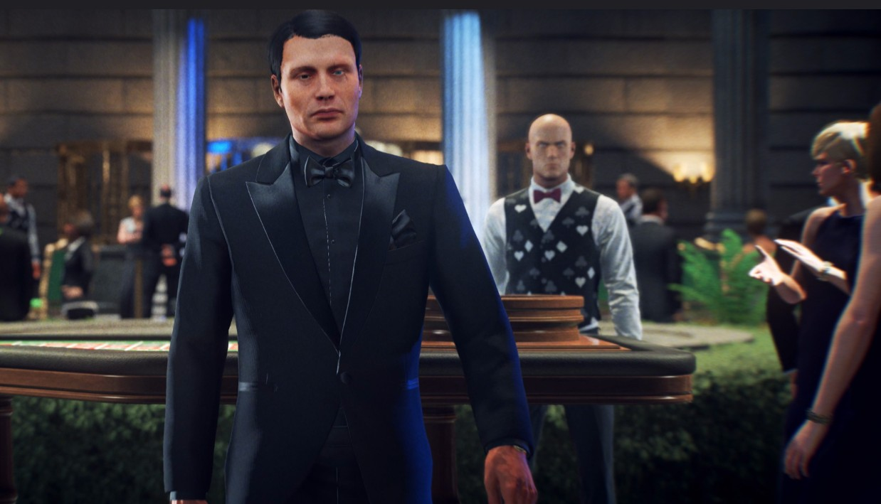 Mężczyzna w czarnym garniturze stoi przy stole w kasynie. Za nim inny mężczyzna w kamizelce i muszce. Ludzie w tle.