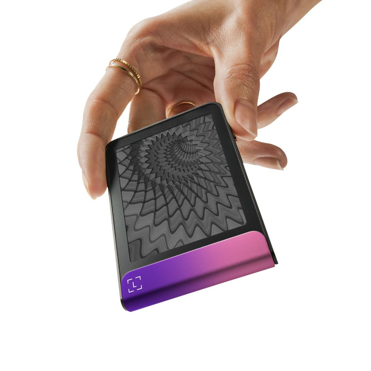 Eine Hand hält ein schwarzes Gerät mit einem pink/lila Farbverlauf am unteren Rand. Der Bildschirm zeigt ein graues Webmuster. Ringe schmücken die Finger.