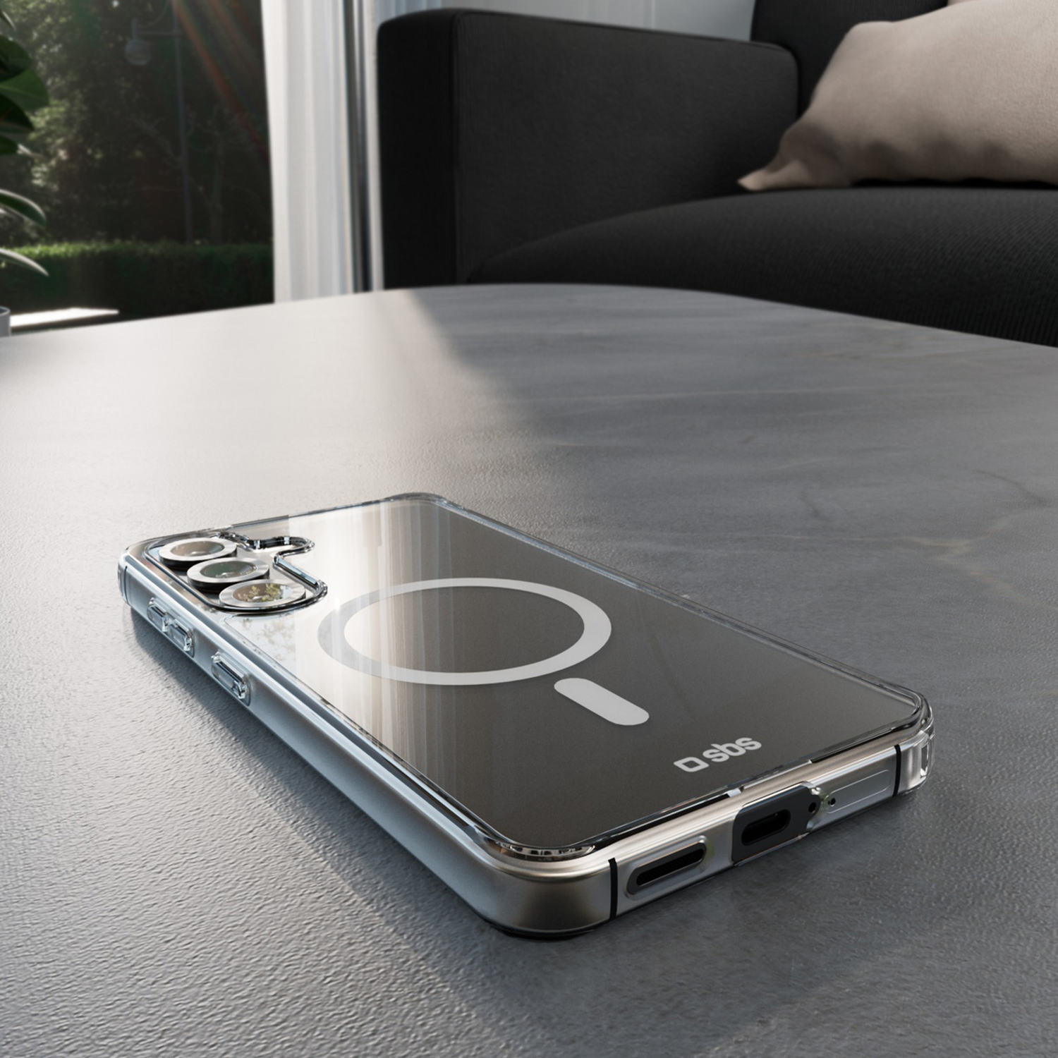 Een doorzichtige telefoonhoes met magnetische ring ligt op een grijze tafel. Op de achtergrond een zwarte bank en een raam.
