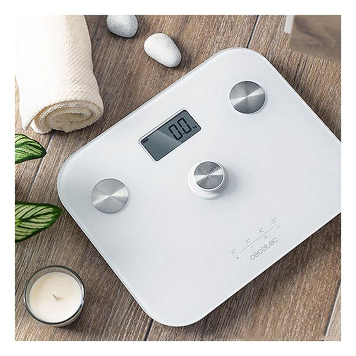 Una báscula digital blanca con pantalla '0.0'. Al lado, una toalla enrollada, piedras, una vela y hojas sobre una superficie de madera.
