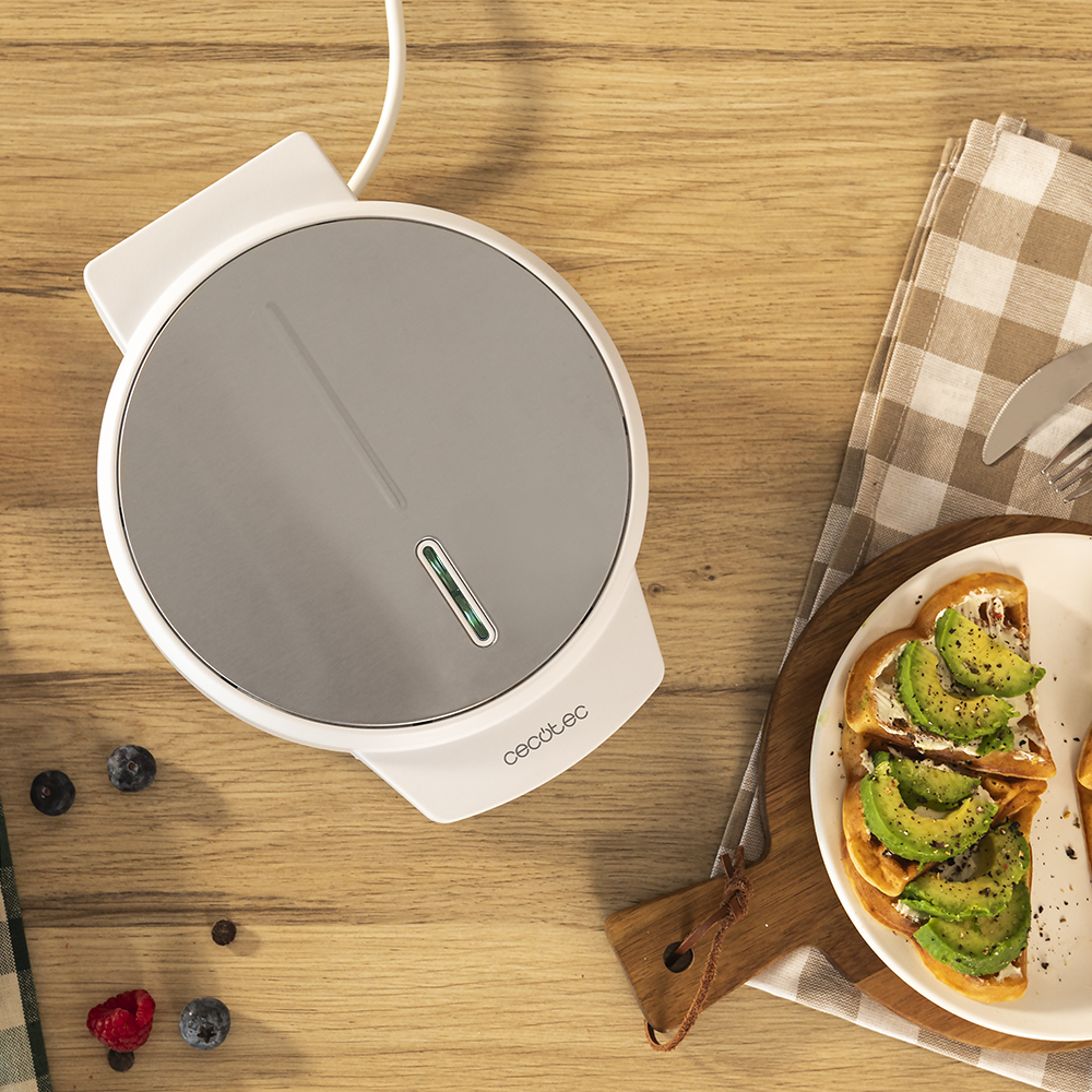 Una gofrera blanca sobre una mesa de madera. Tostadas de aguacate en un plato. Bayas y un paño a cuadros también están en la mesa.