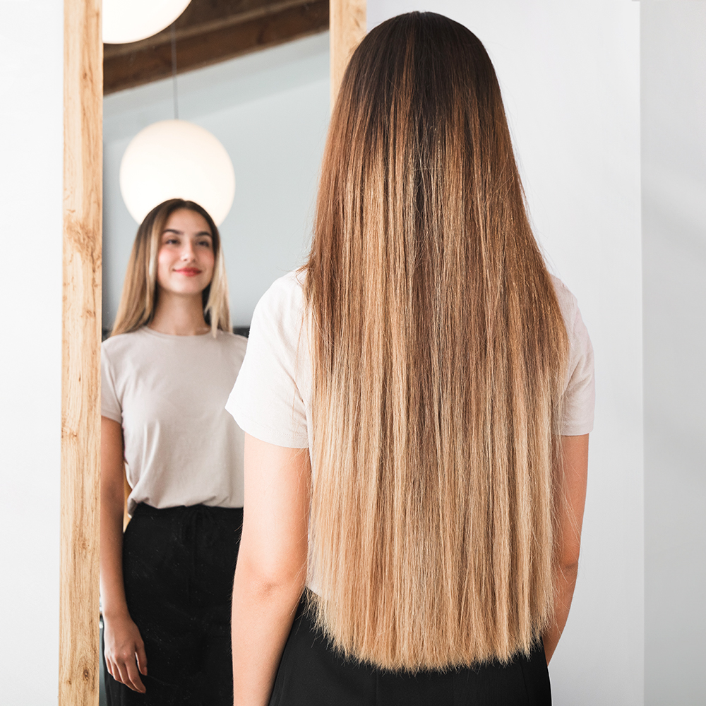 Mujer con pelo largo mirándose en un espejo. El espejo refleja su rostro sonriente. Lleva una camisa clara y una falda negra.