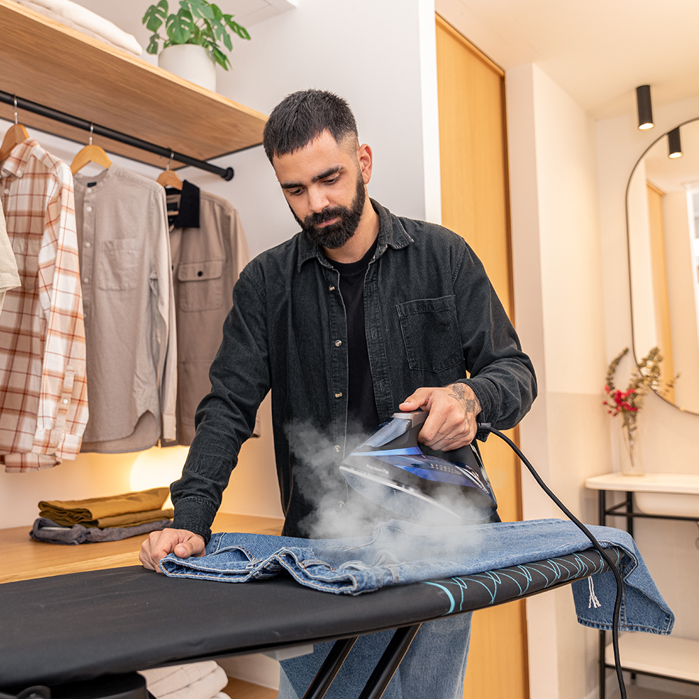 Un hombre plancha unos vaqueros sobre una tabla de planchar. El vapor sale de la plancha. La ropa cuelga al fondo. Un espejo y flores son visibles.