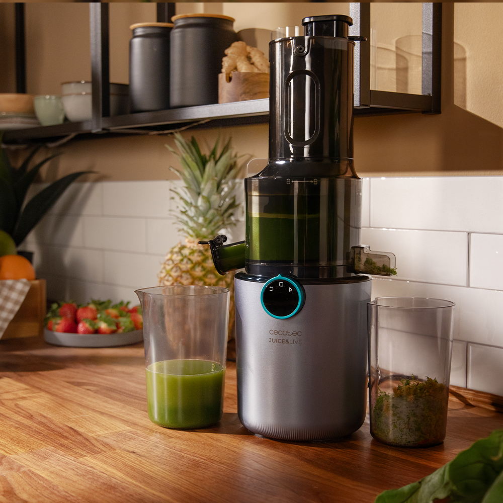 Una exprimidora está sobre una encimera de madera. Un vaso de jugo verde y un recipiente con pulpa. Se ven piña y fresas.