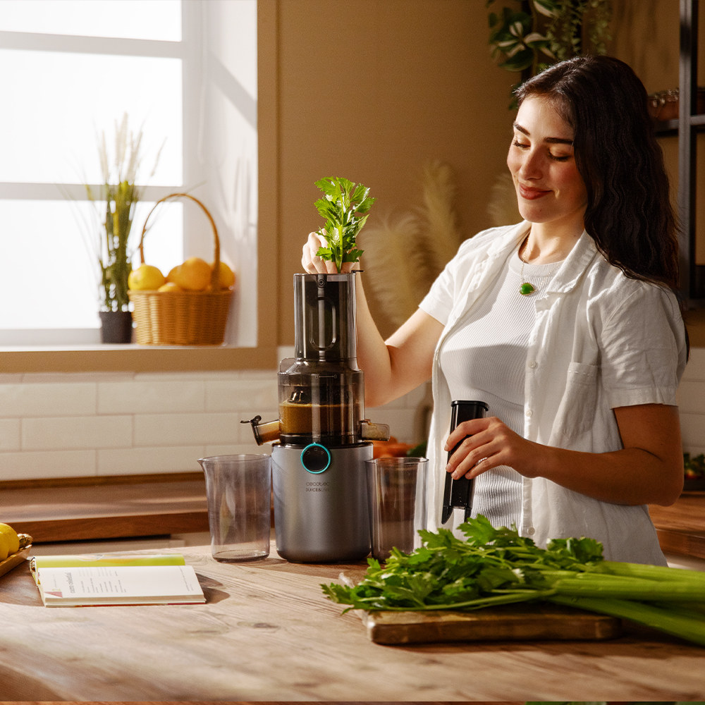 Mujer agregando apio a una juguera. Escena de cocina con ventana, cesta de naranjas y mesa de madera con ingredientes.