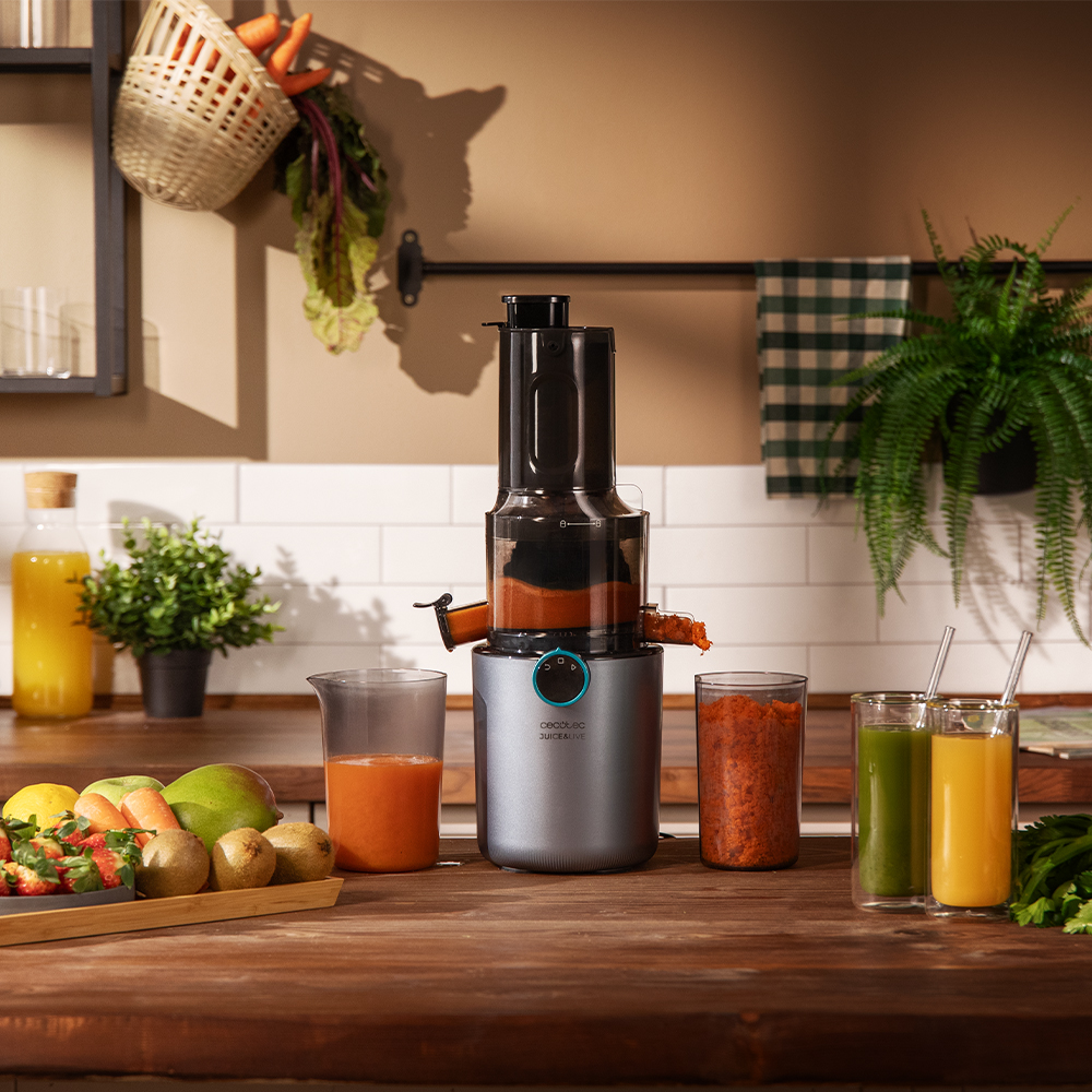 Exprimidor con frutas y verduras. El exprimidor es plateado y negro. Jugos en vasos. Frutas en una bandeja. Verduras en una cesta.