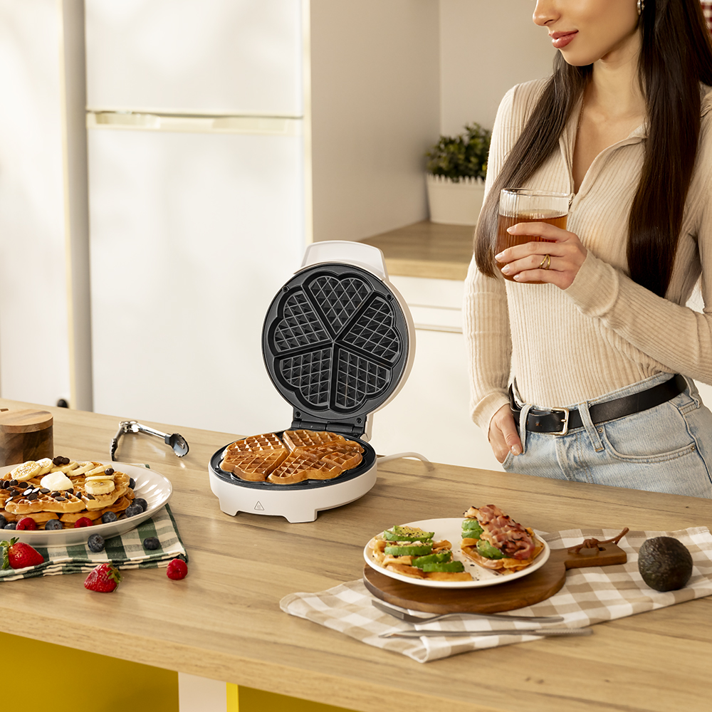 Una mujer sostiene té cerca de una máquina de hacer gofres. Los gofres están en la máquina y en platos con ingredientes como frutas y aguacate.