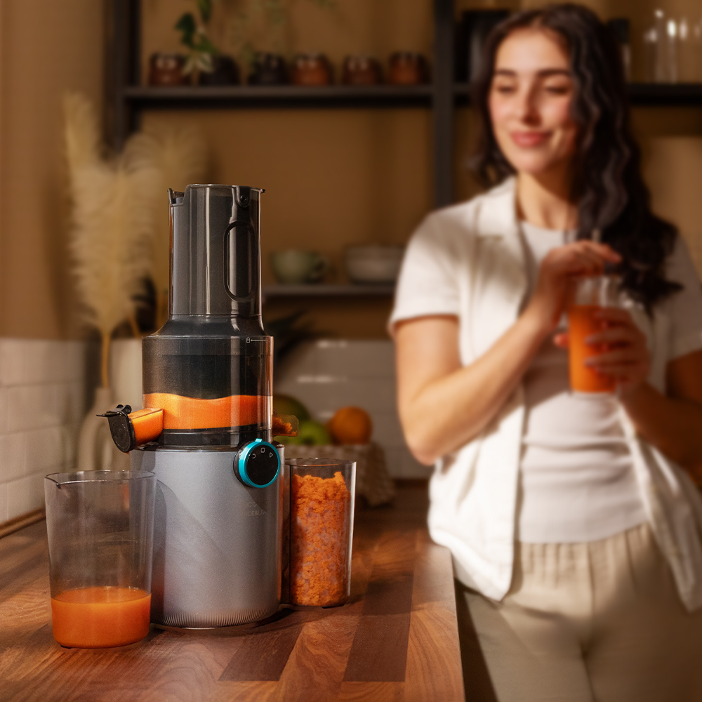 Una mujer está de pie cerca de una exprimidora, sosteniendo zumo. La exprimidora es gris y naranja. Dos vasos contienen zumo y pulpa.