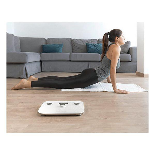 Mujer en ropa deportiva haciendo yoga sobre una esterilla. Una báscula está frente a ella. Un sofá gris está en el fondo.