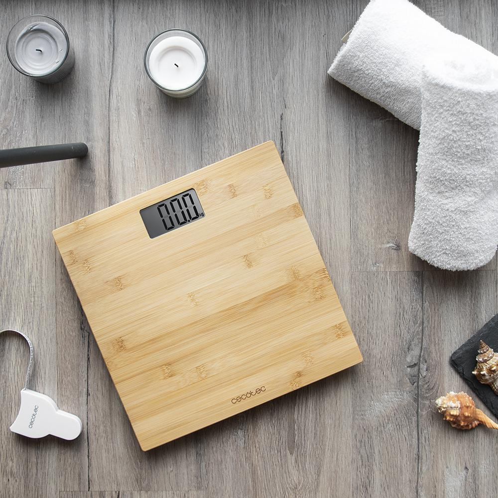 Una báscula de madera con pantalla '0.00', velas, toallas blancas, conchas y una cinta métrica sobre madera.
