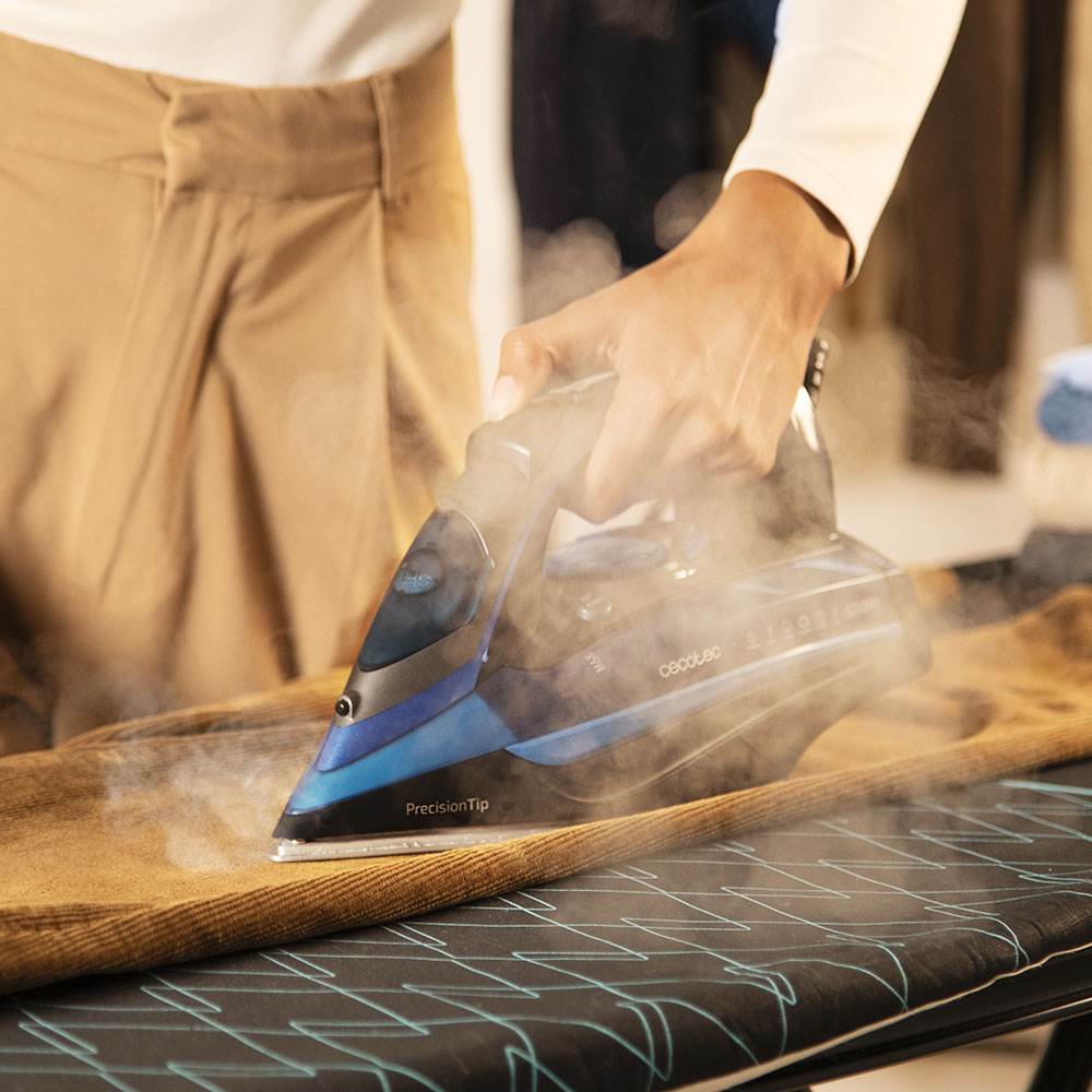 Una persona planchando pantalones marrones con una plancha azul, vapor ascendente. Tabla de planchar con diseño negro y azul en primer plano.