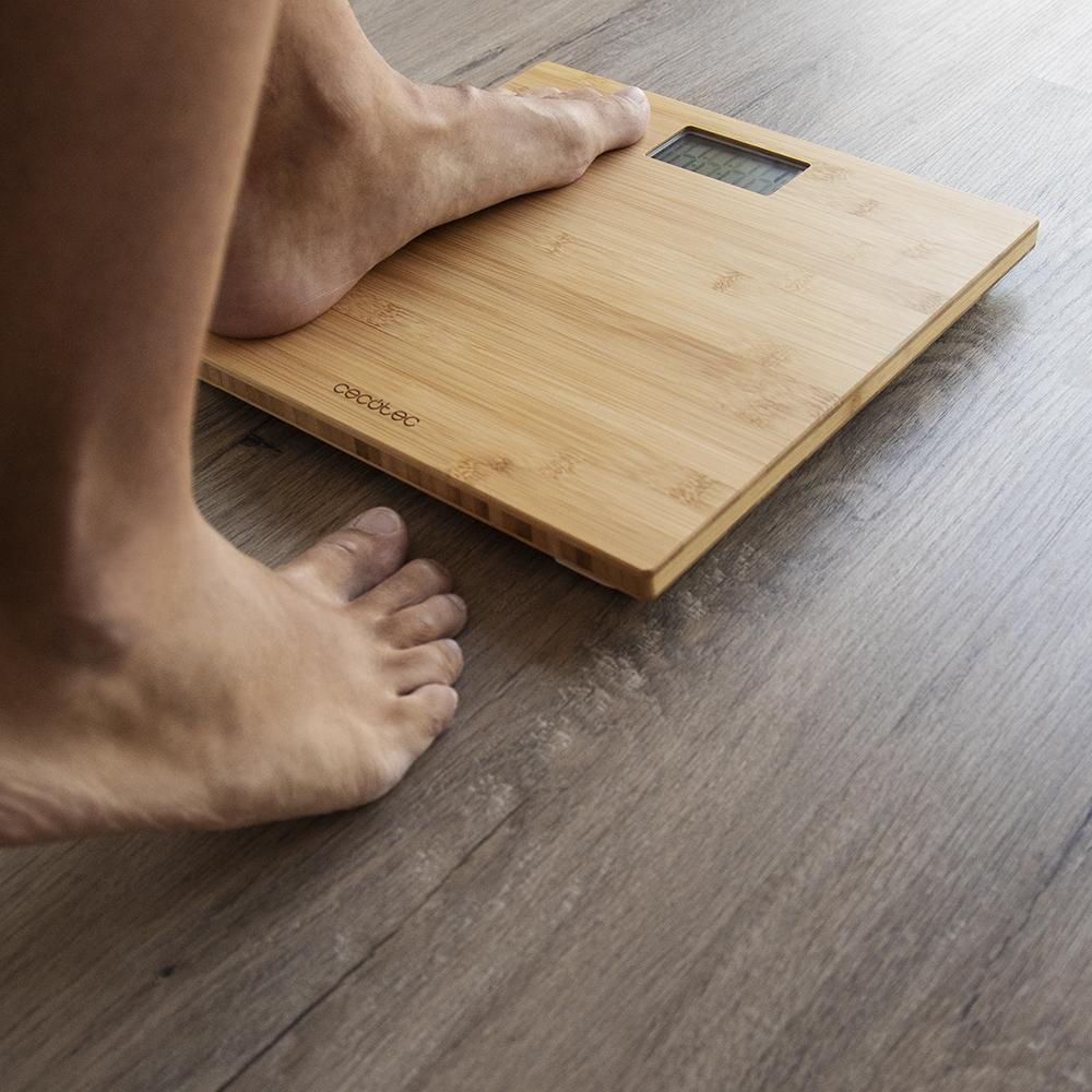 Pies de una persona en una báscula de bambú sobre suelo de madera. La báscula muestra números. Un pie está en la báscula, el otro al lado.