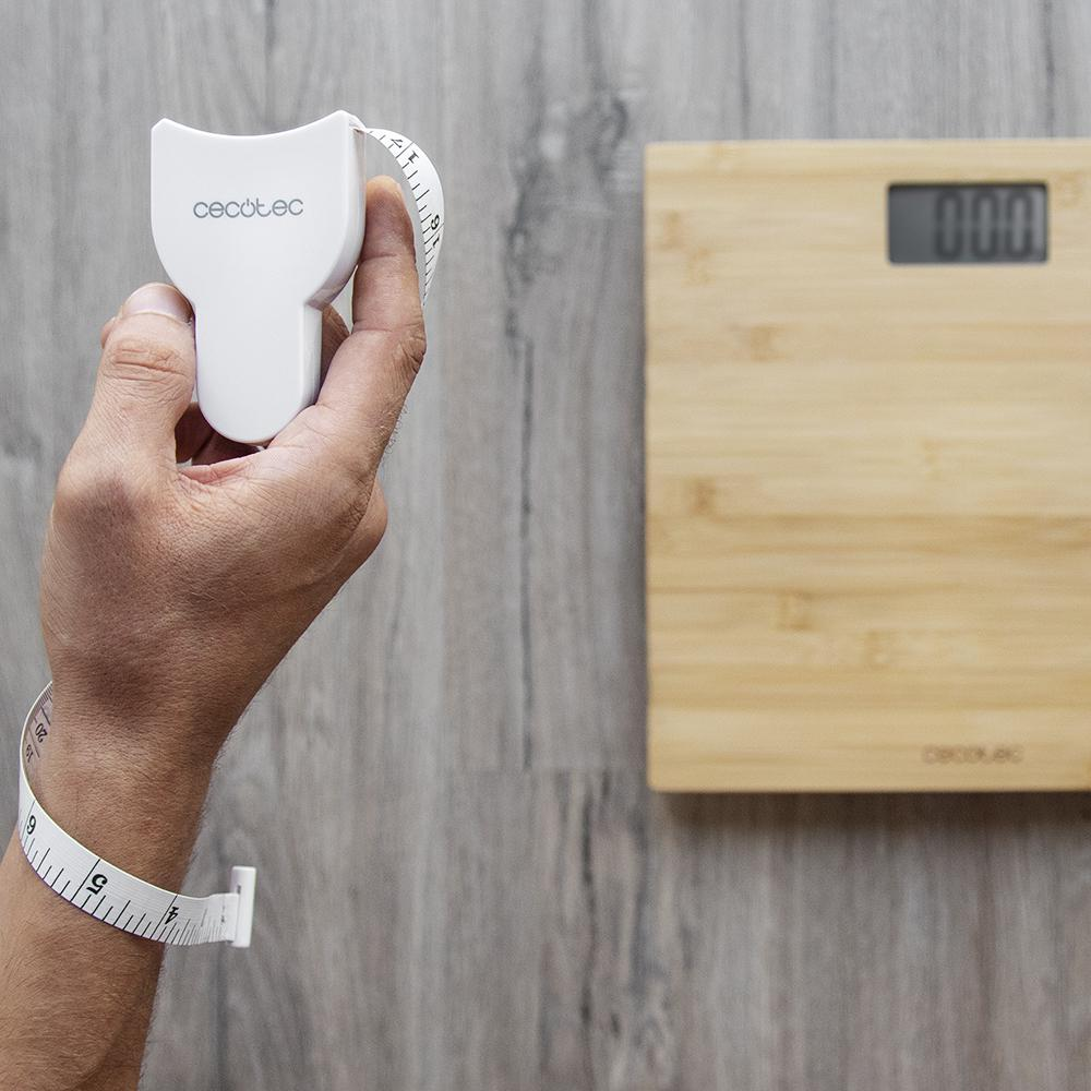 Una mano sostiene una cinta métrica blanca para el cuerpo. Una cinta métrica envuelve la muñeca. Una báscula de madera está al fondo.