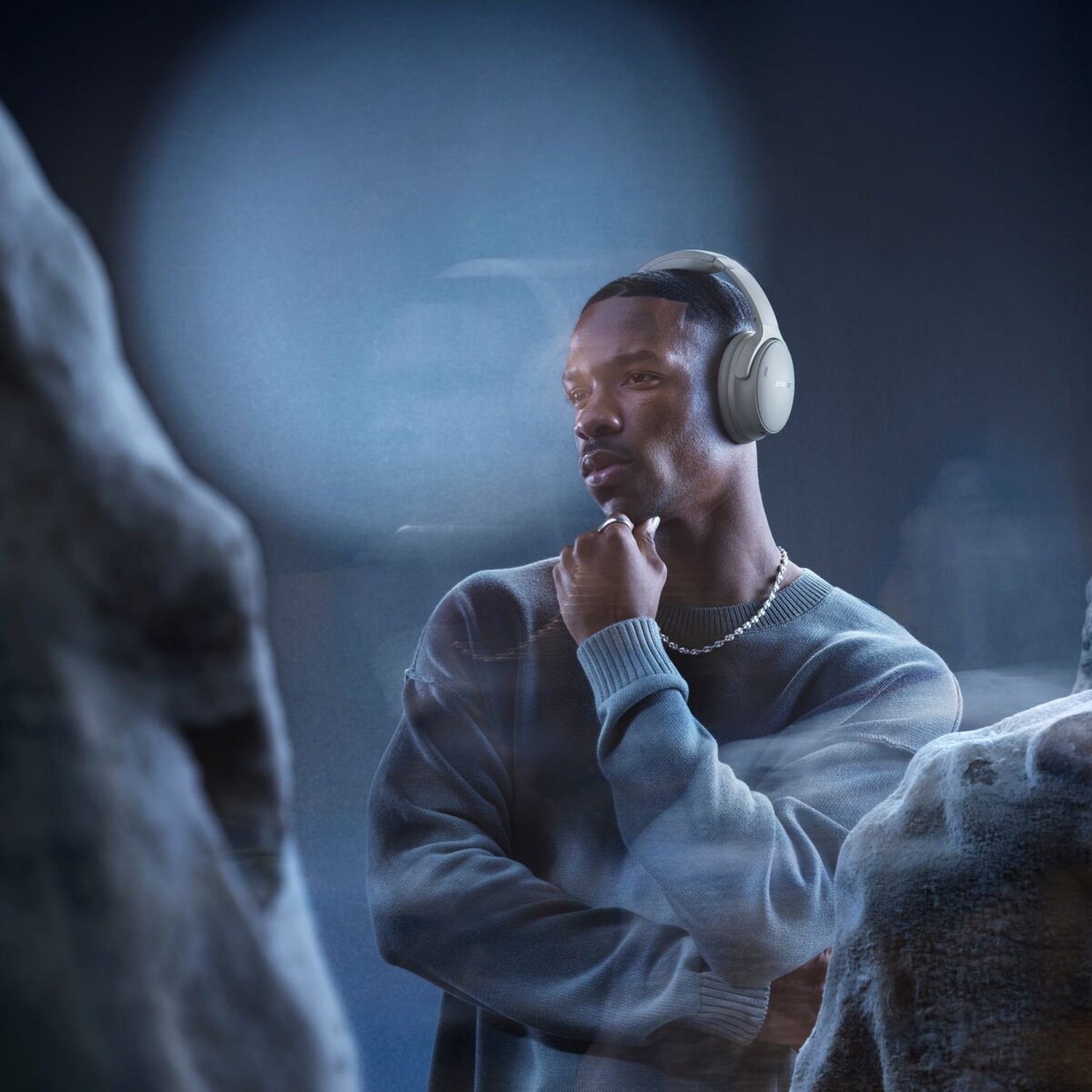 A Black man wears headphones, a light blue sweater, and a silver necklace. He is looking off to the side, touching his chin.