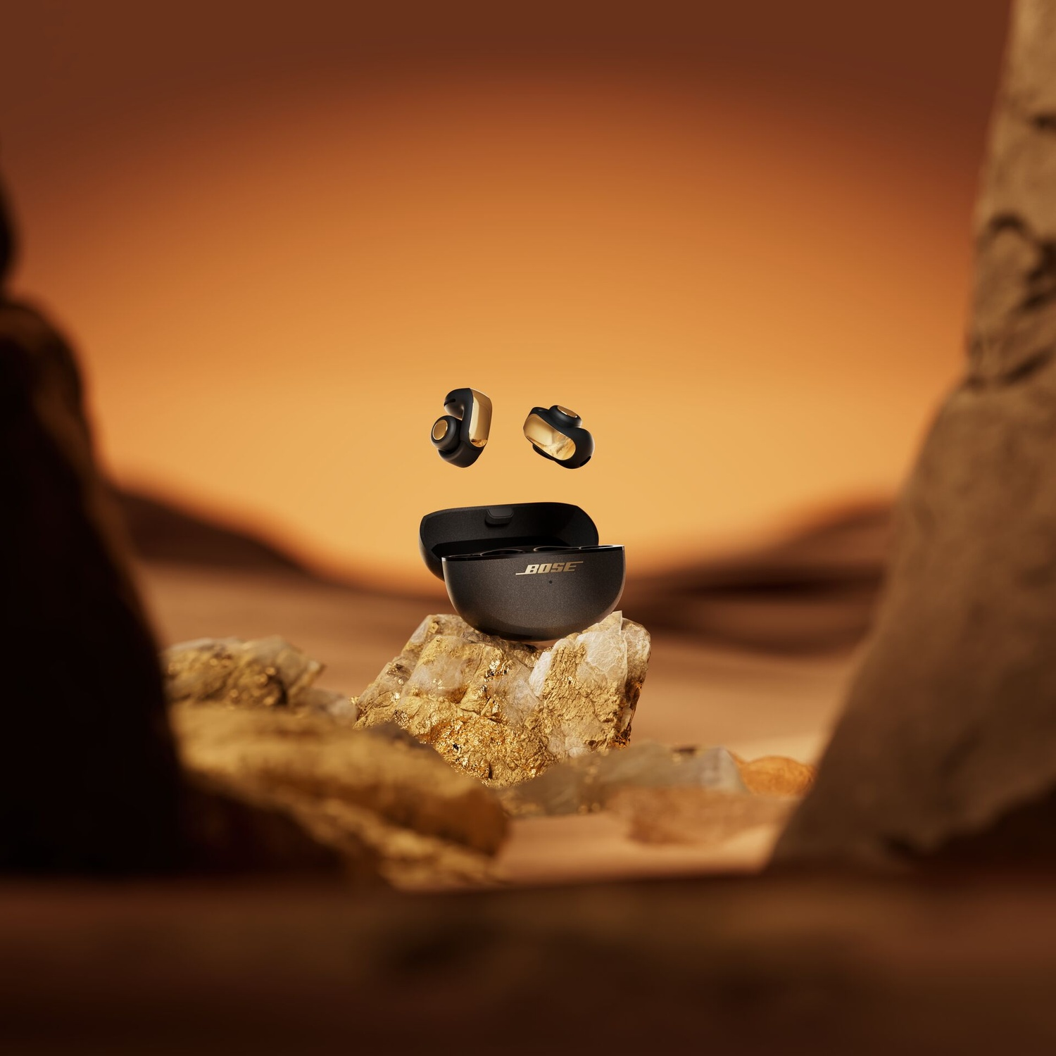 Earbuds and case floating over stone. Gold and black earbuds. Orange and brown background. Bose logo on the case.