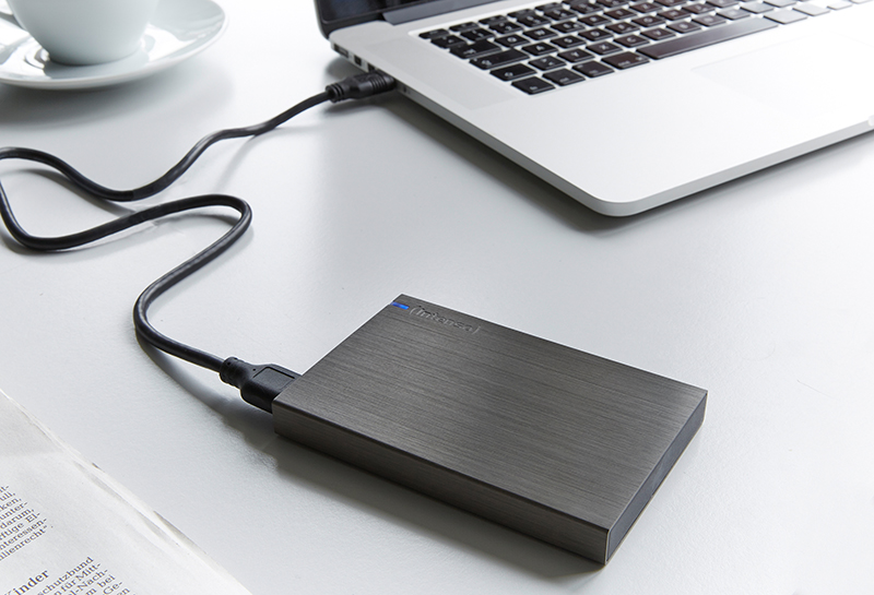 Un ordinateur portable argenté connecté à un disque dur externe gris via un câble noir. Une tasse blanche à gauche.