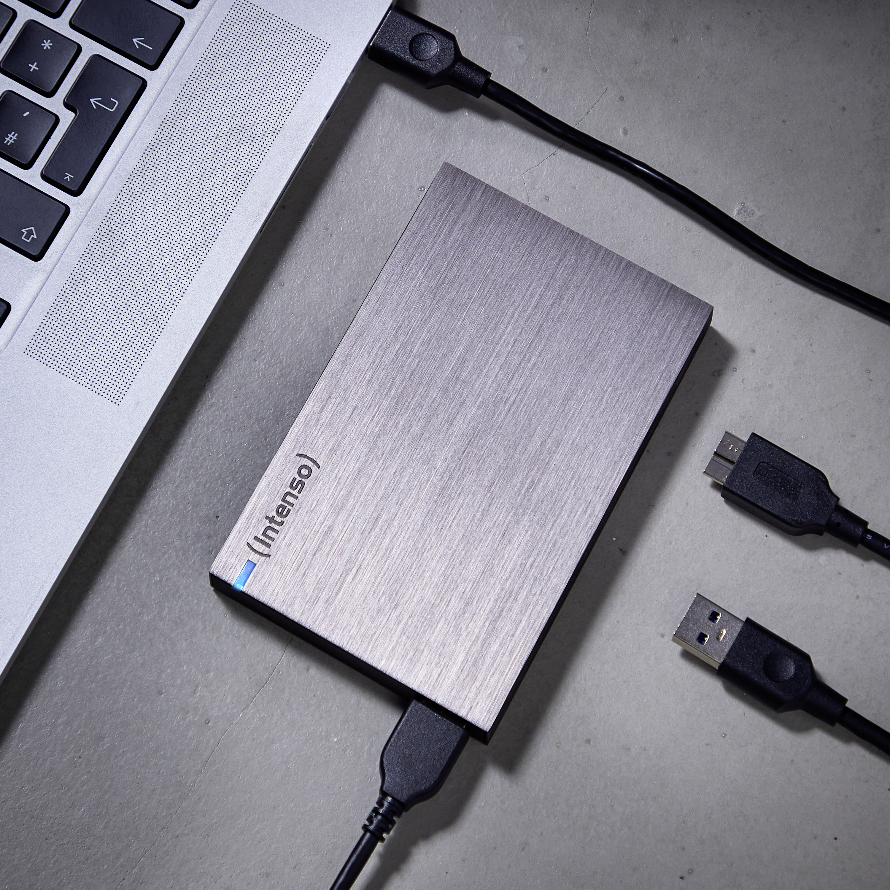 Disque dur externe argenté étiqueté (Intenso) connecté à un ordinateur portable. Des câbles USB sont également visibles sur une surface en béton.
