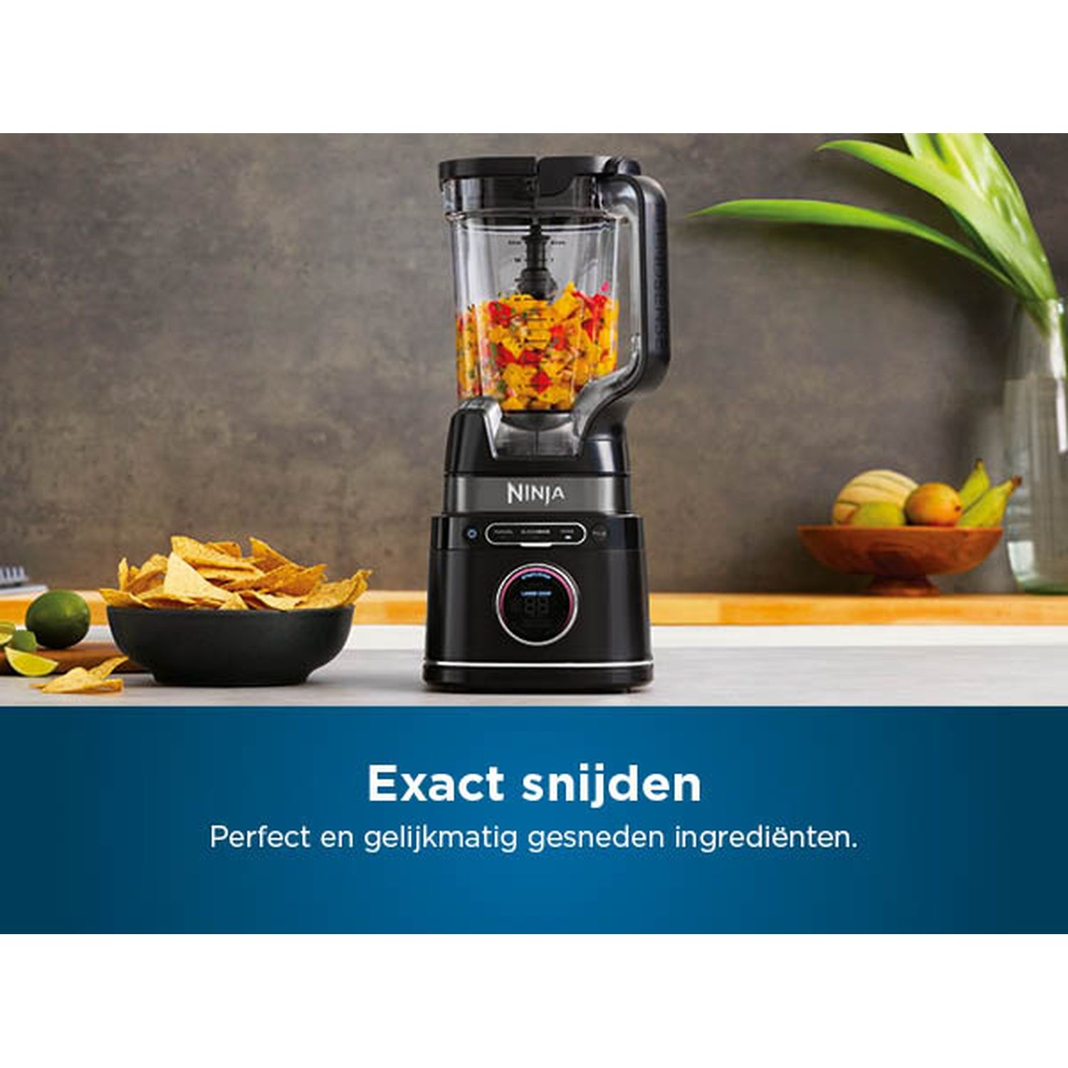Un blender Ninja noir avec des légumes hachés à l'intérieur. Des chips de tortilla dans un bol noir à gauche. Des fruits dans un bol en bois derrière.