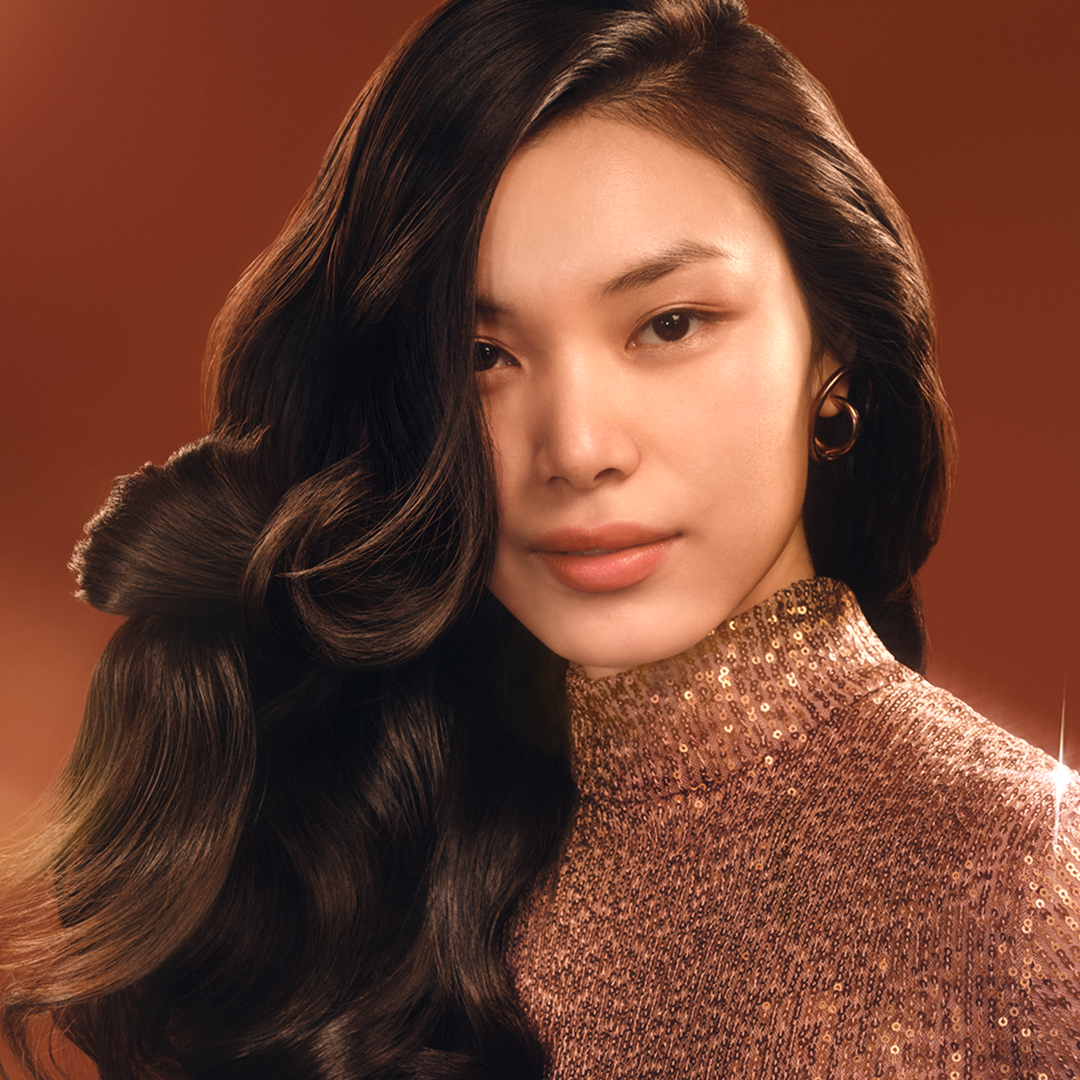 Kobieta z ciemnymi, falowanymi włosami i złotym golfem z cekinami patrzy w kamerę. Nosi kolczyki w kształcie kółek. Brązowe tło.