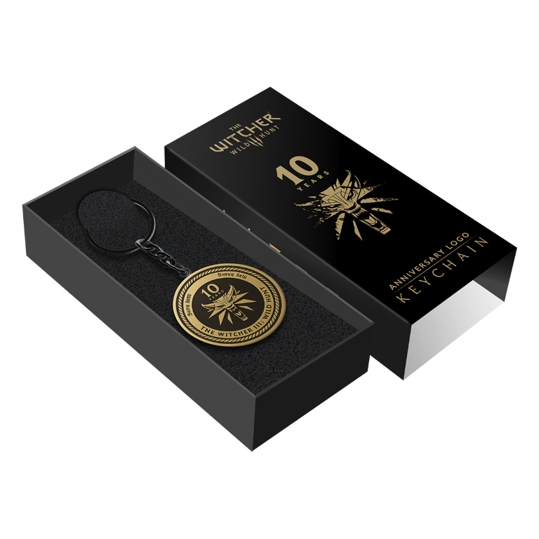 Złoty brelok Witcher III na 10-lecie w czarnym pudełku. Pudełko i brelok zawierają logo gry i tekst.