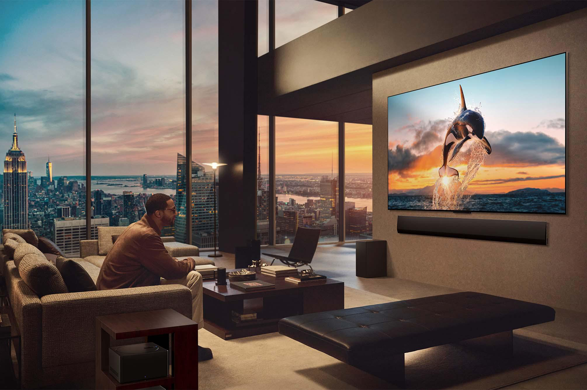 Ein Mann sitzt in einem Luxusraum und betrachtet einen Fernseher, der einen springenden Orca zeigt. Stadtblick durch Fenster.