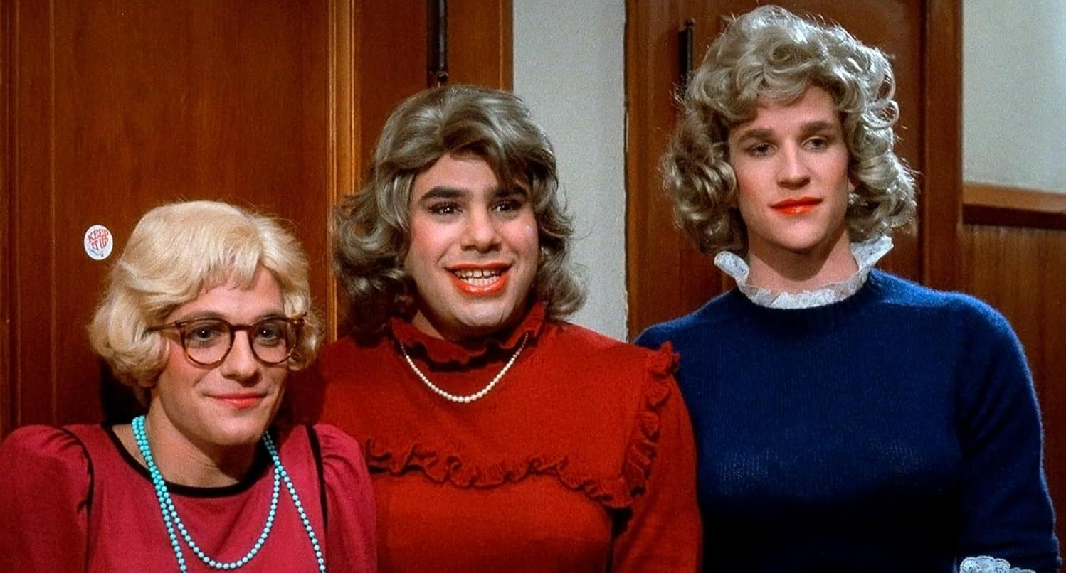 Drei Männer in Frauenkleidern, mit Perücken und Make-up. Einer trägt eine Brille und ein rotes Outfit, die anderen Kleider, Perlen und blaue Pullover.