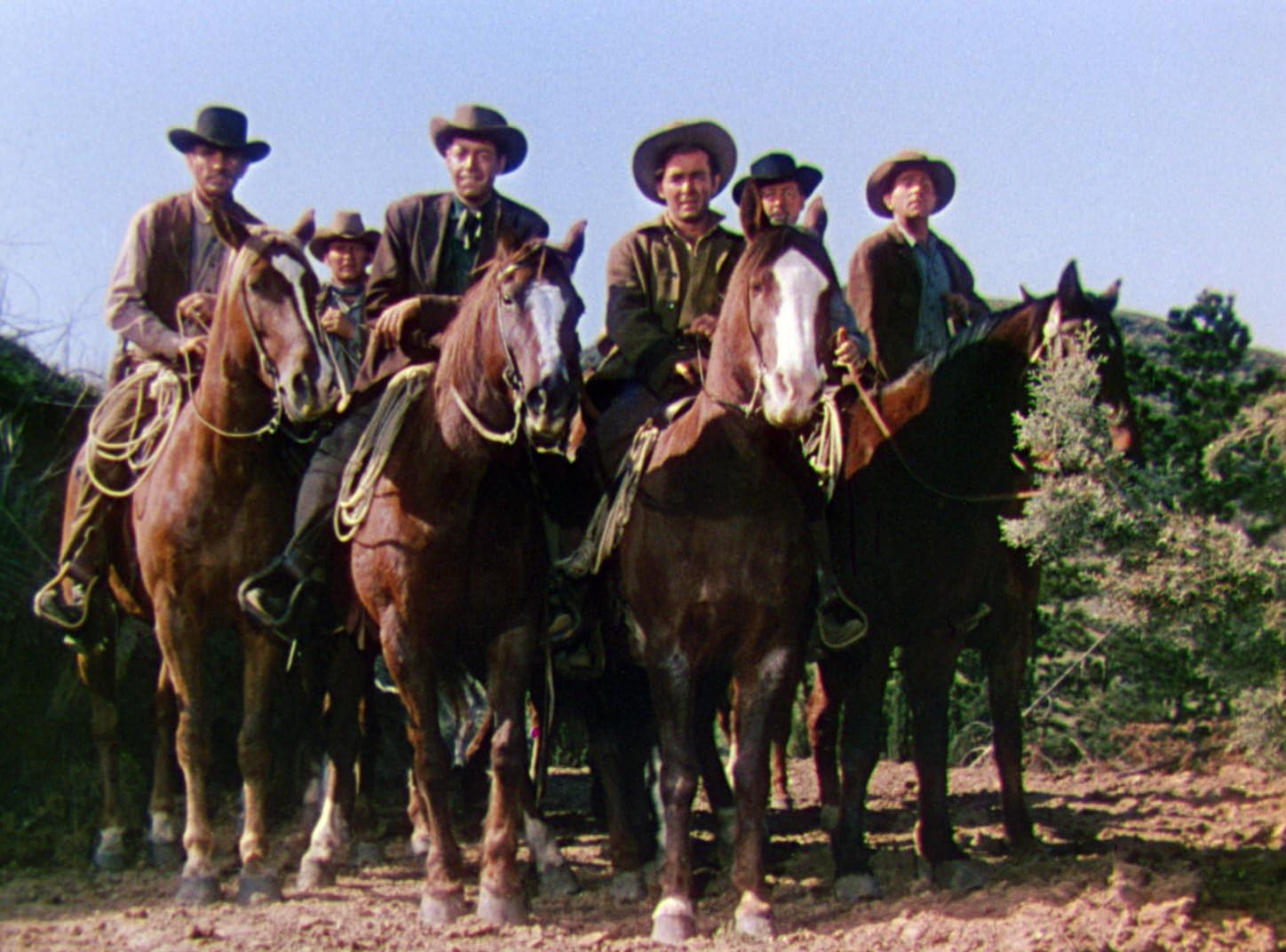 Sieben Männer auf Pferden mit Cowboyhüten. Pferde sind braun und schwarz. Die Szene spielt in einer wüstenartigen Umgebung mit Büschen.